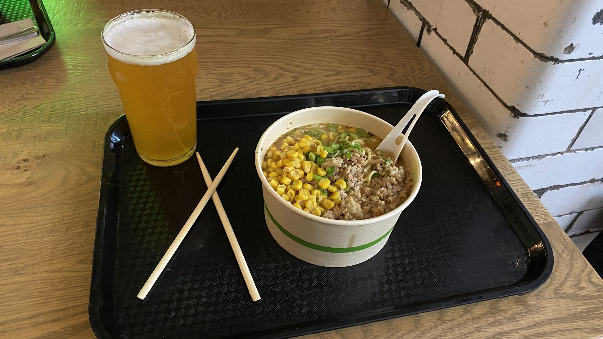 A ramen bowl topped with corn, ground pork and scallions with chopsticks and a glass of beer next to it
