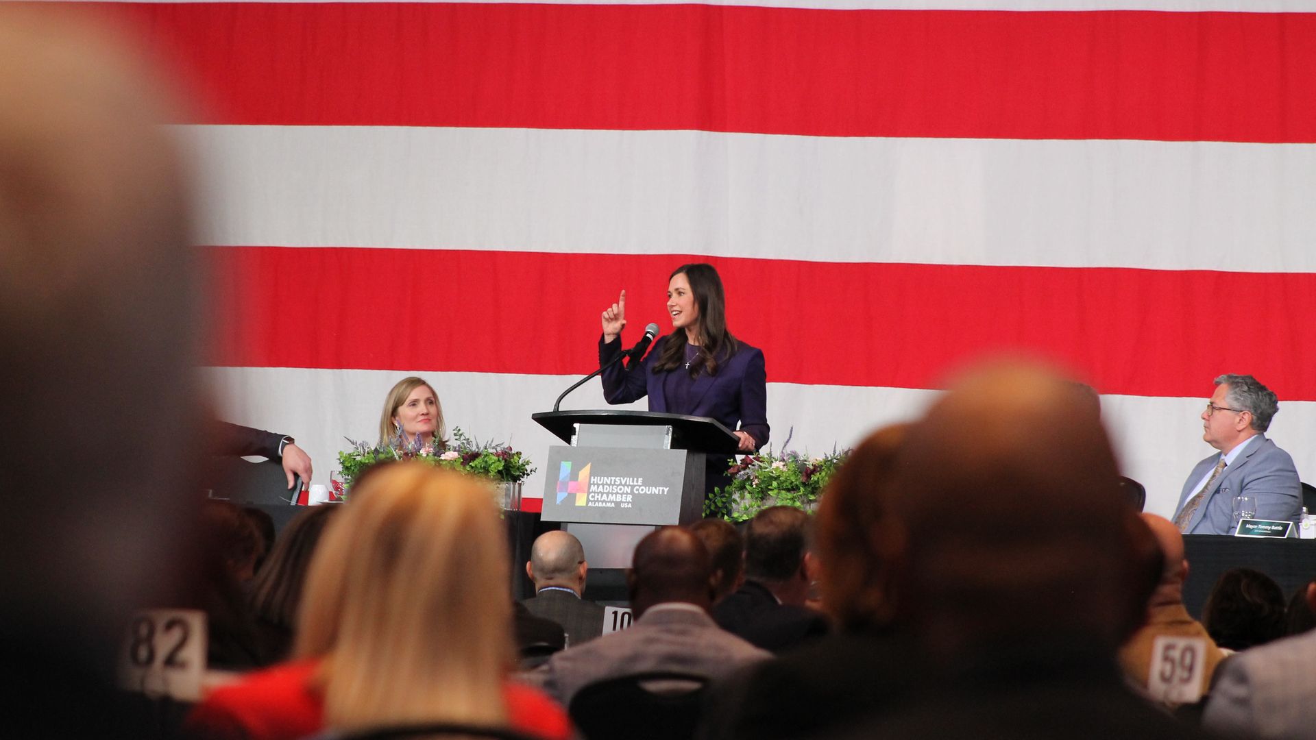 A woman in a dark suit speaks at a podium with the Huntsville Madison County Chamber logo, in front of a large American flag, addressing an audience with seated panelists nearby.