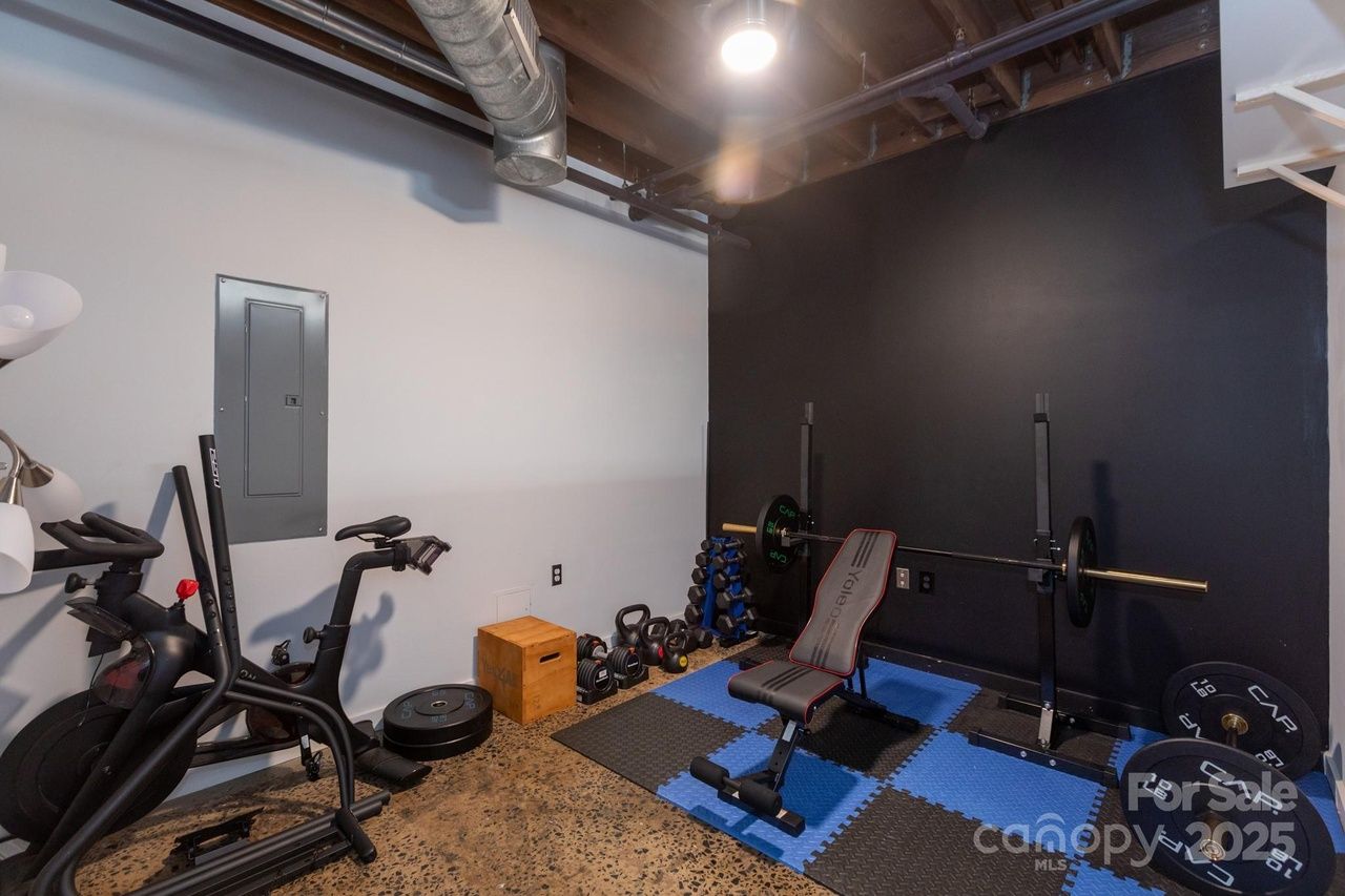 Home gym with black and blue foam floor mats, black adjustable weight bench, barbell rack with weights, kettlebells, dumbbells, and stationary exercise bike against white and black walls.