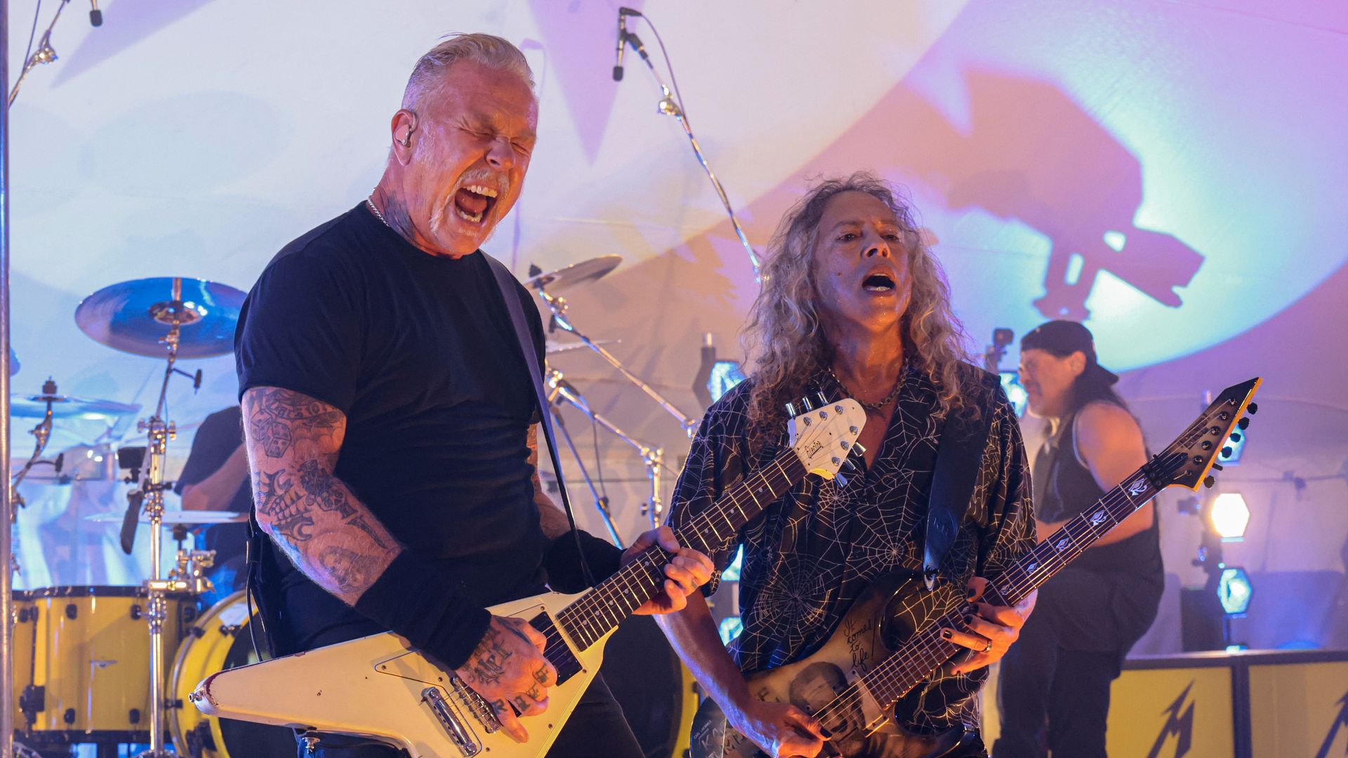 Rock musicians on a lit stage: a tattooed, gray-haired man screams into a microphone while playing a white Flying V; beside him, a long-haired bassist in a patterned shirt plays a dark bass.