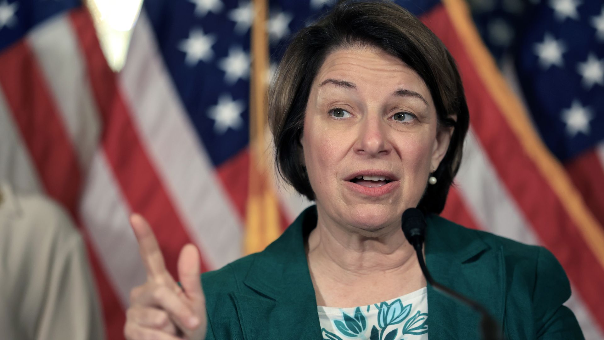 Woman in green jacket speaking at a microphone with American flags in the blurred background, gesturing with her right hand raised and finger pointing up.