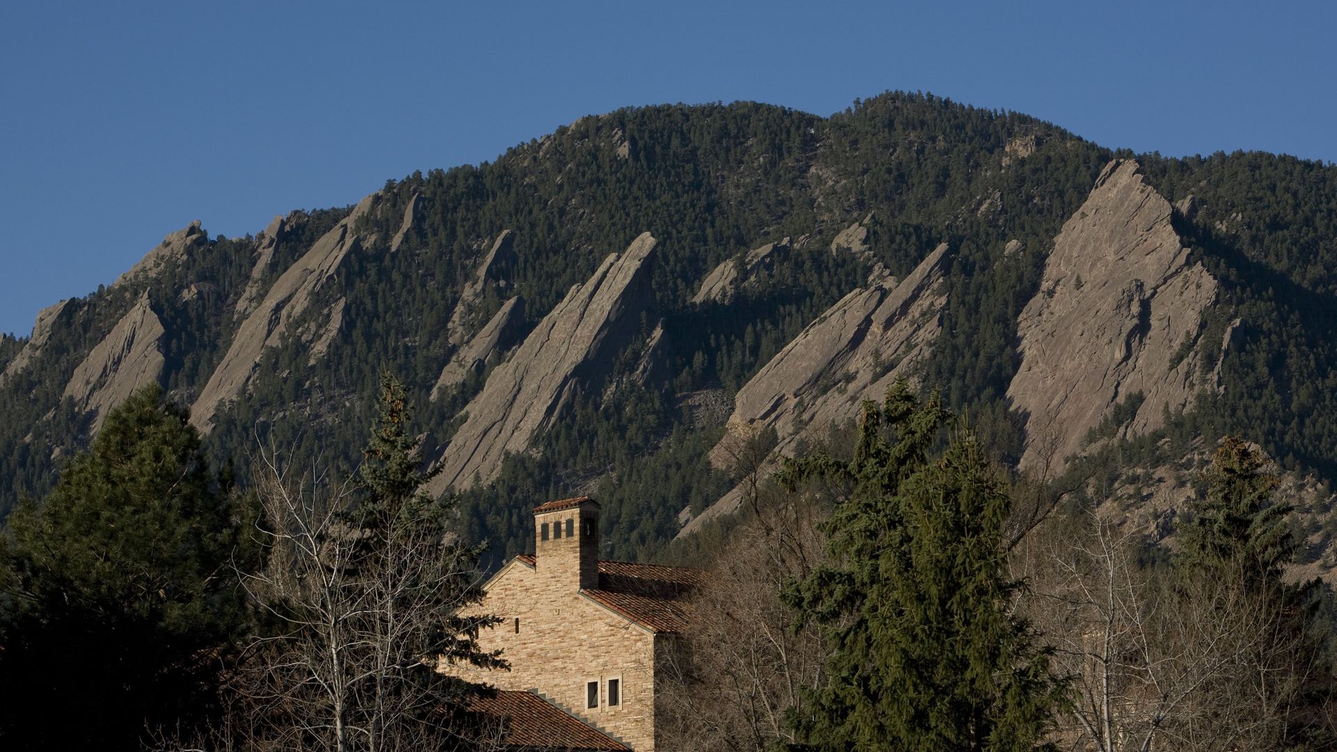 Rocky, tree-covered mountain ridge under a clear blue sky, with diagonal stone spires. In the foreground a stone building with a red-tiled roof and a chimney, surrounded by bare trees.