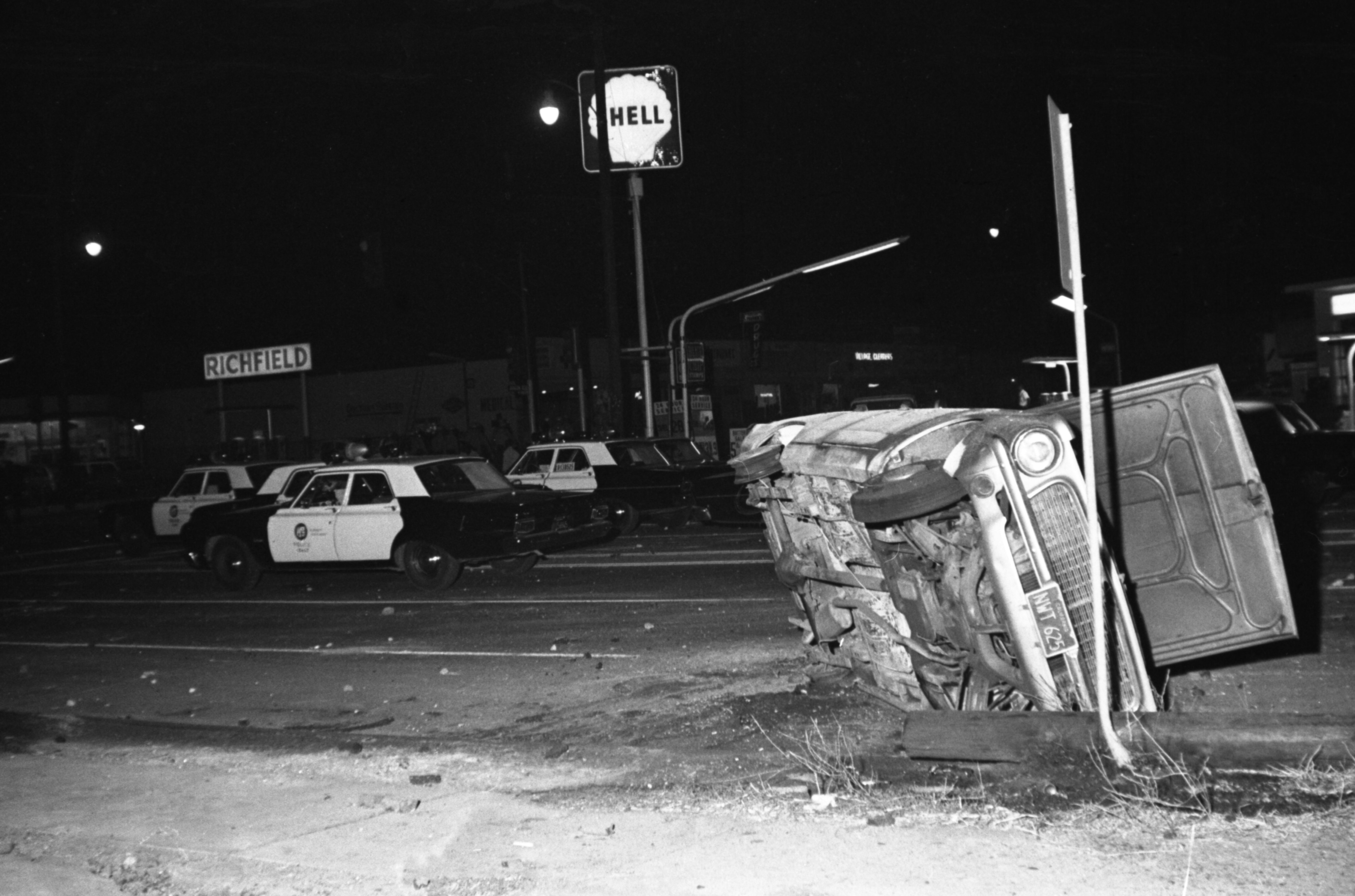 Black and white night photo showing an overturned car next to a tilted street sign with three parked police cars nearby and a partially visible illuminated sign reading Shell.