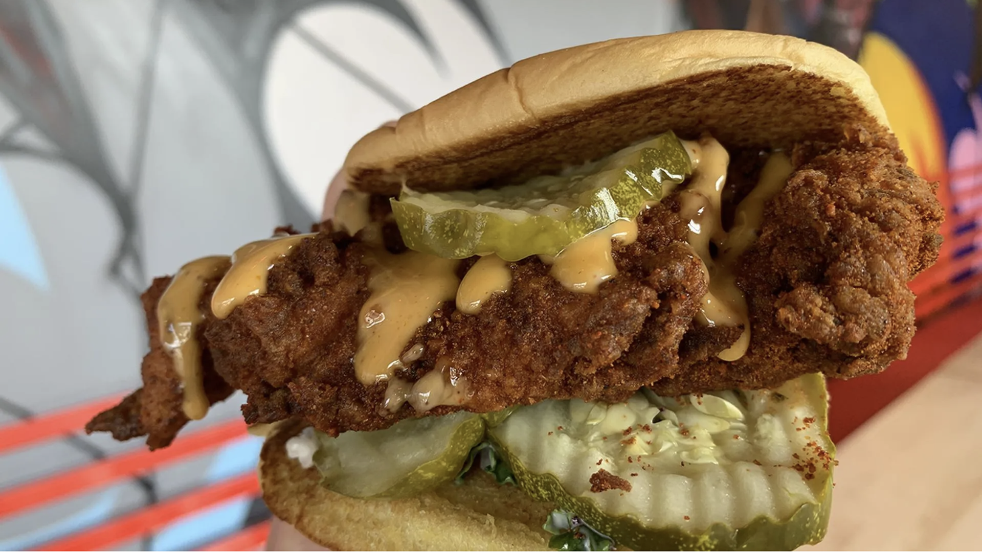 Close-up of a large fried chicken sandwich: crispy fillet, creamy orange sauce, and pickle slices in a soft bun, with shredded lettuce and more pickles underneath.