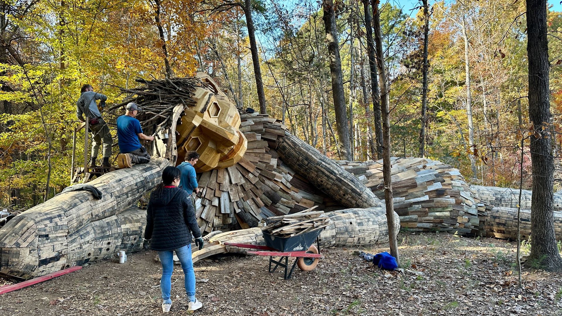 Four people working on a large wooden sculpture of a reclining giant with a wooden face and twig hair in a forest with autumn foliage.