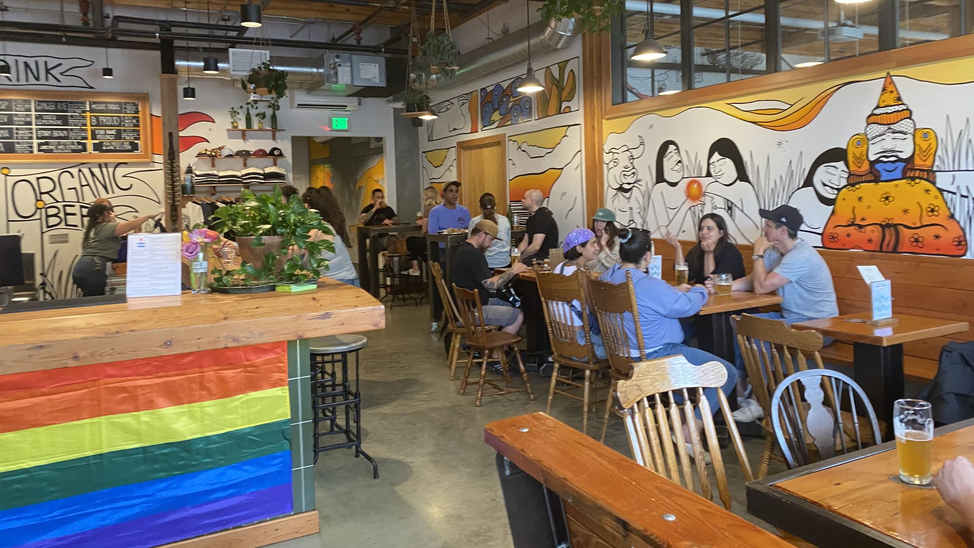 A room with tables and people sitting at them, with murals on the walls and a pride flag at the end of a bar.