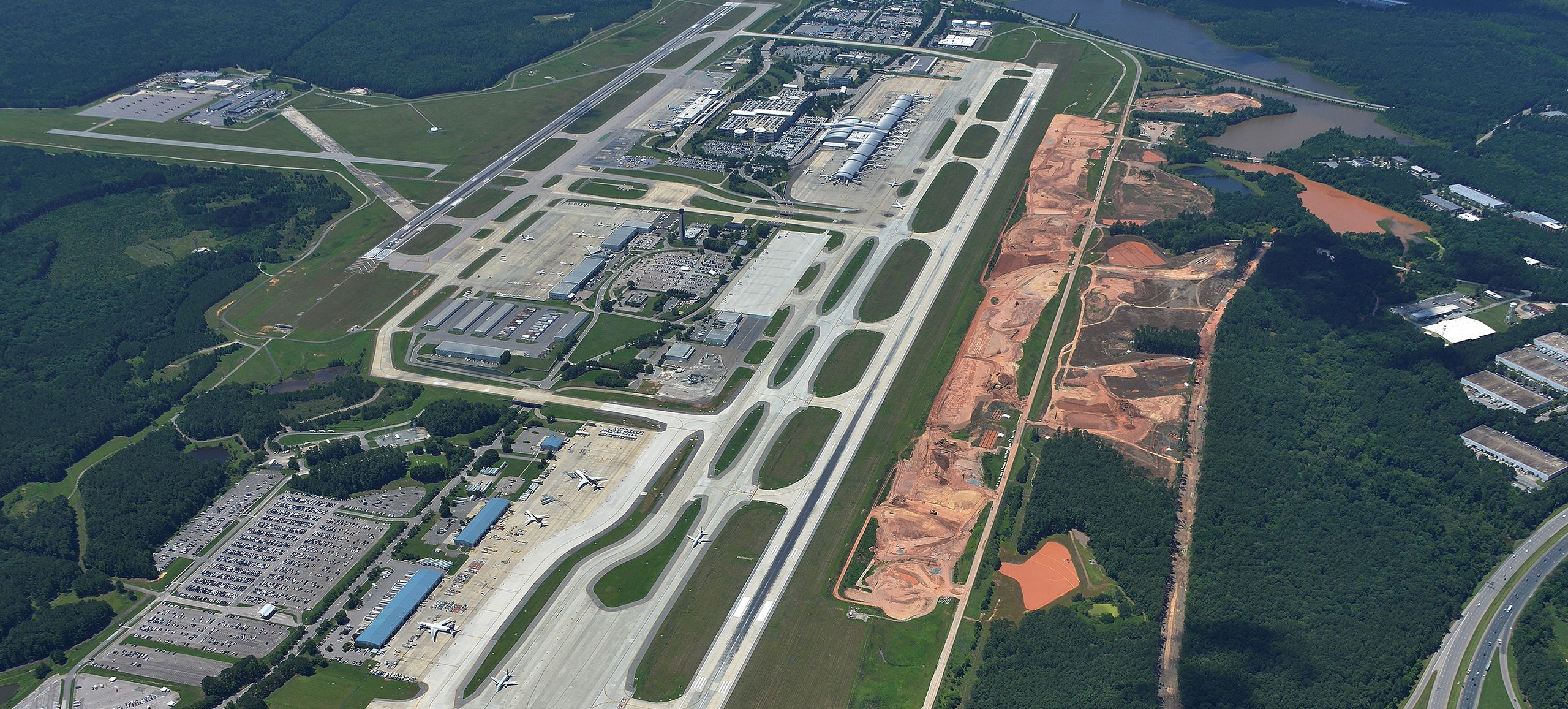 Aerial view of an airport with multiple runways, terminals, parked airplanes, surrounding greenery, and an adjacent construction site with exposed red soil and dirt roads.
