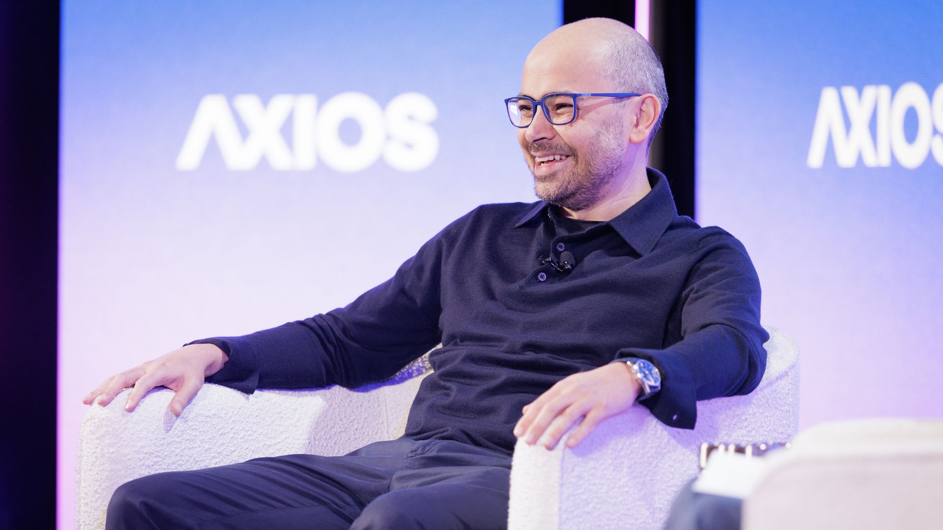 Smiling bald man with glasses wearing a black shirt, seated in a white armchair on stage with blue background displaying "AXIOS" logo.