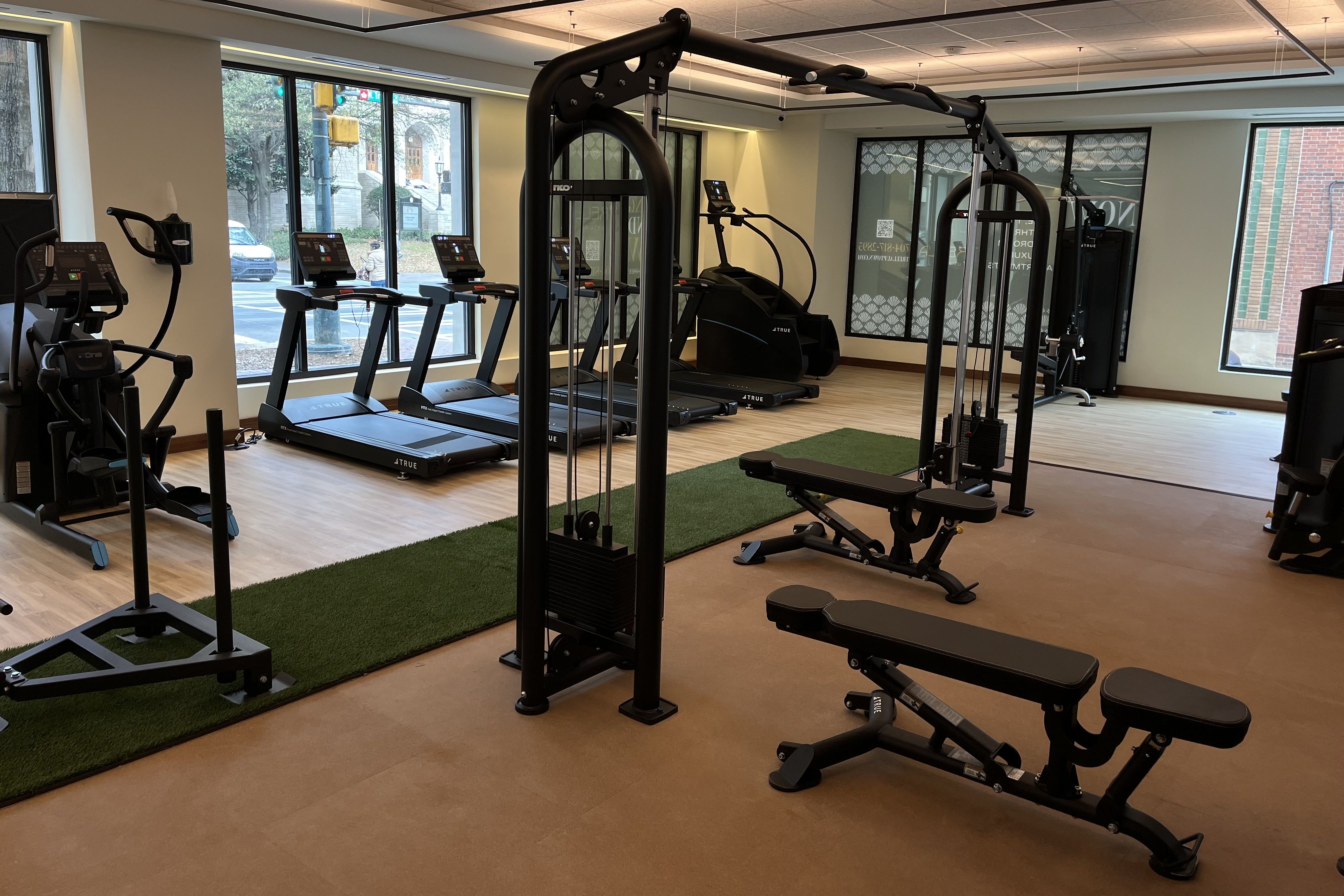 Indoor gym with black workout benches, cable machine, treadmills, elliptical, and wood flooring with green turf sections near large windows showing an outdoor street scene.