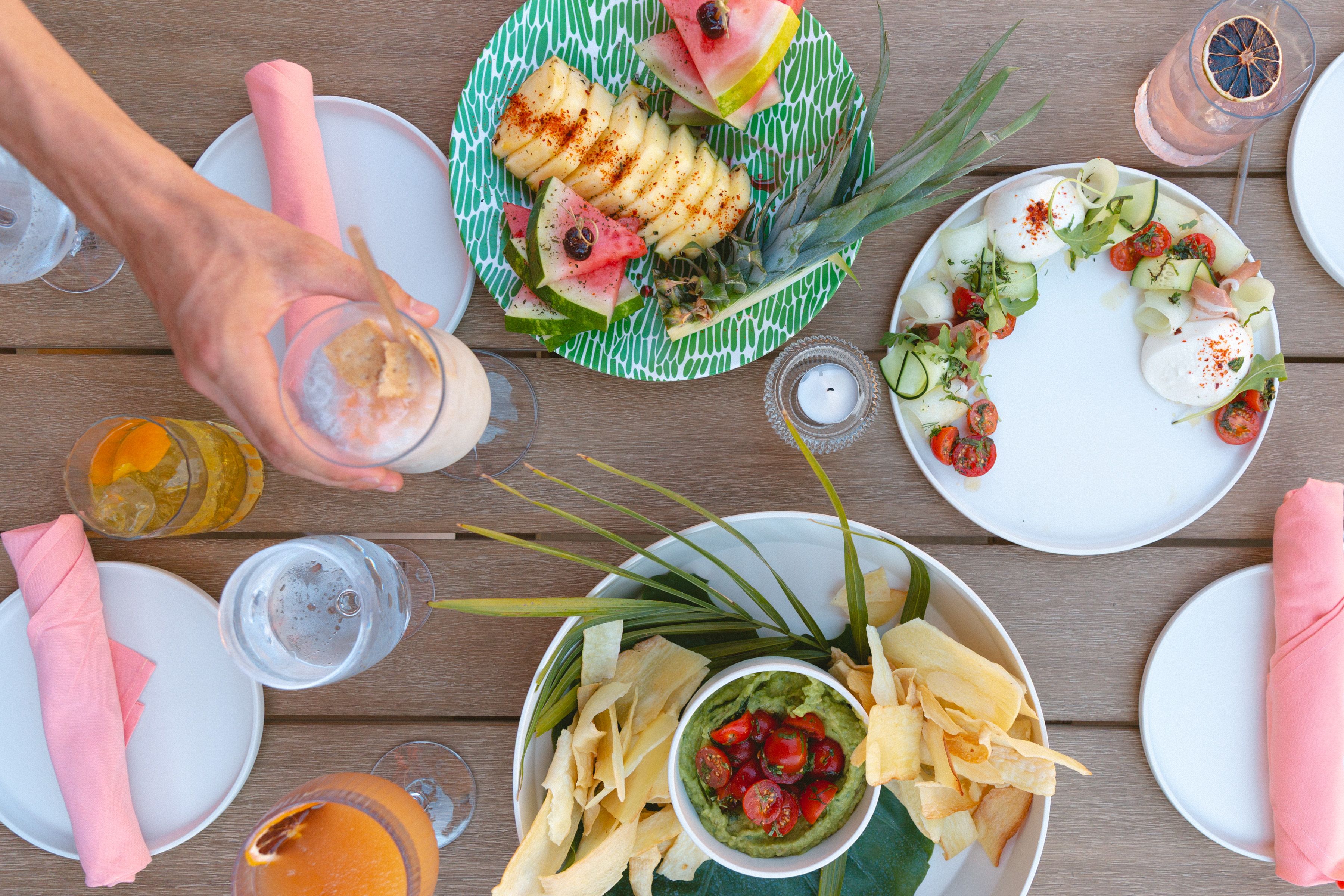Table with tropical fruit plate, guacamole with chips, a salad with cherry tomatoes and mozzarella, various drinks including a cocktail with orange slice, and pink napkins.