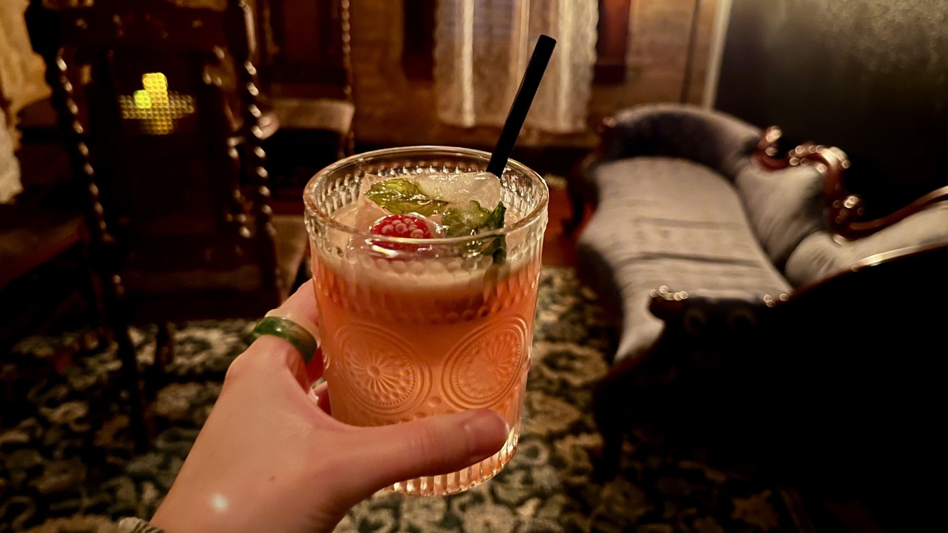 A pink-colored cocktail with a large ice cube with mint and raspberry, with Victorian furniture in the background.