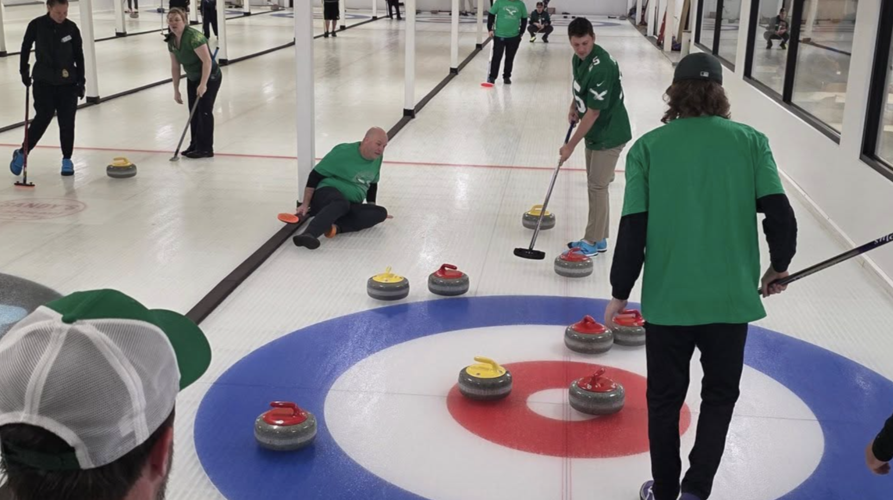 Indoor curling game with several players in green shirts, one sitting on the ice. Multiple red and yellow curling stones near a target with red, white, and blue rings.