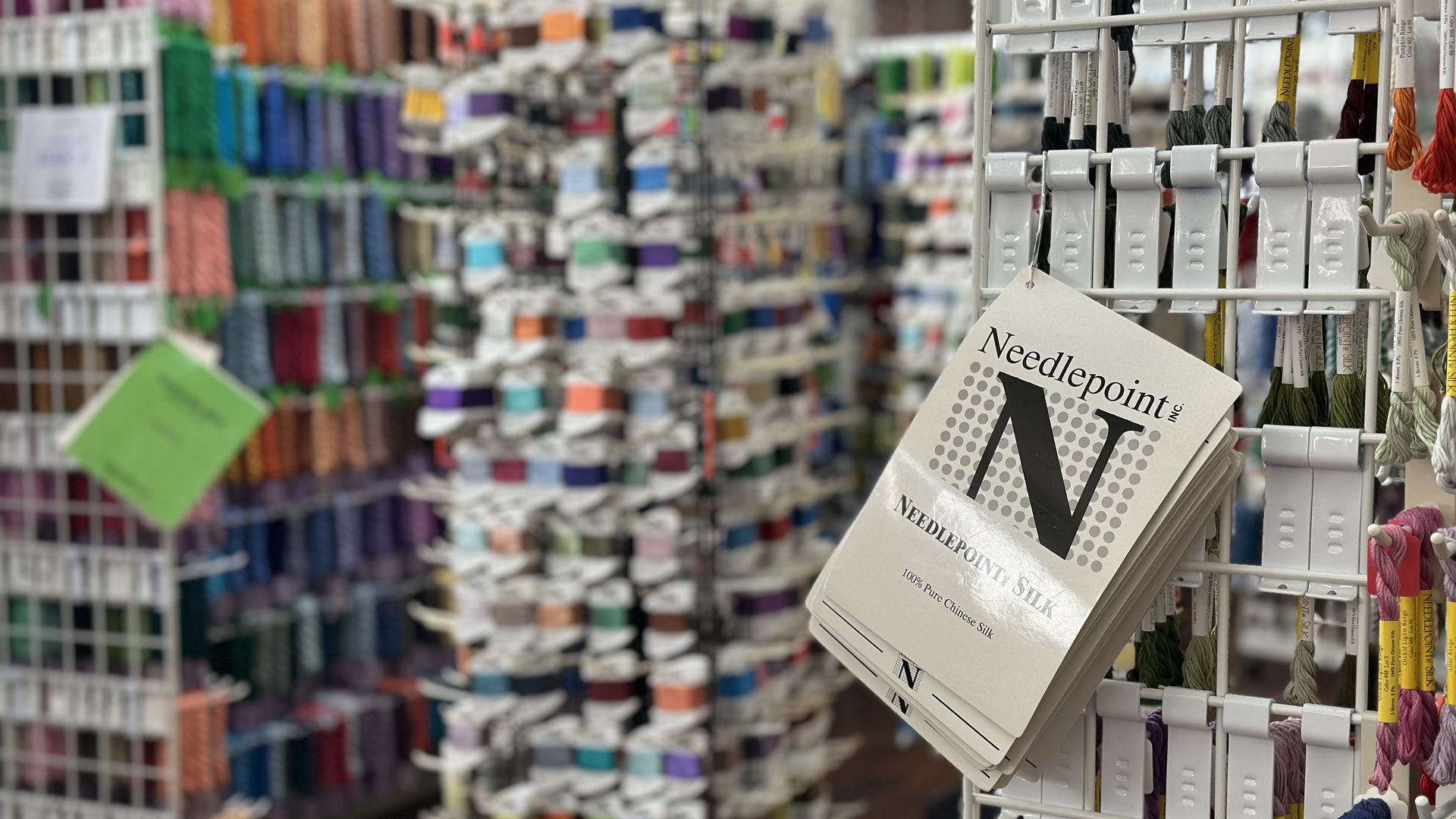 Inside a craft store, a rack of multicolored embroidery floss spools; a tilted card reads "Needlepoint Silk" with a large "N" on the front, while blurred thread shelves appear in the background.