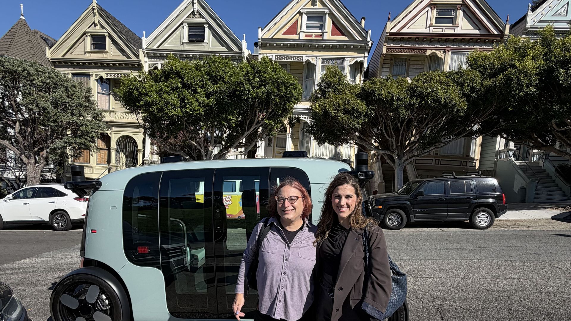 Two smiling women stand beside a light blue compact car on a sunny street with ornate row houses and green trees, in front of San Francisco's Painted Ladies.