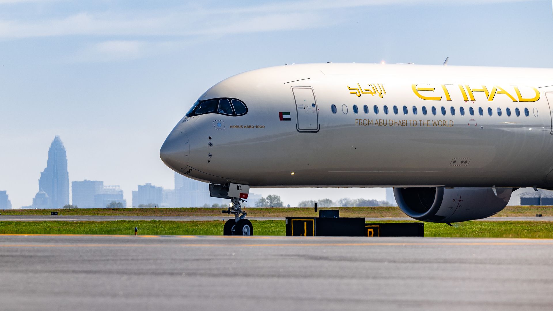 A white Etihad Airbus A350-1000 on a taxiway, with gold ETIHAD lettering and a UAE flag near the nose; the slogan FROM ABU DHABI TO THE WORLD visible, Charlotte skyline and blue sky in the background.