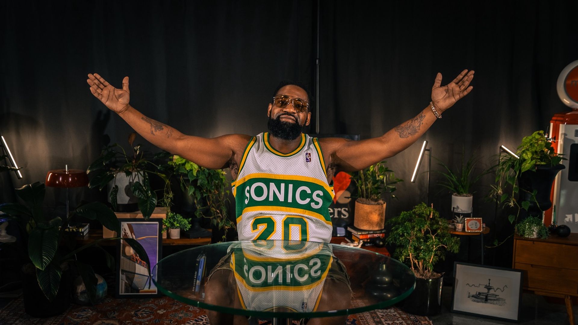 A man wearing a Sonics #20 jersey with his hands outstretched while sitting down in front of plants.