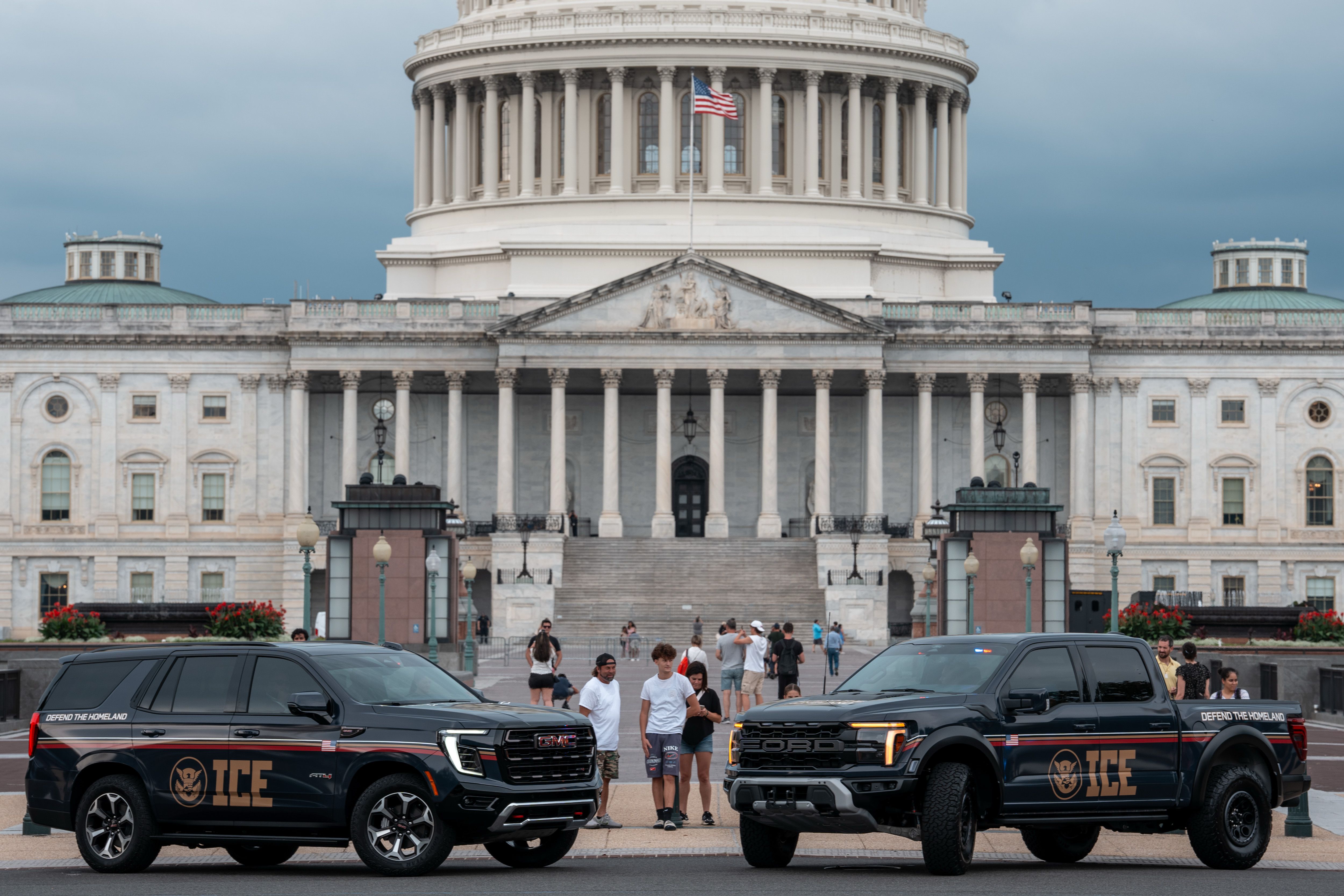 Two black ICE vehicles, an SUV and a pickup truck, parked in front of the U.S. Capitol building with people walking nearby under a cloudy sky.