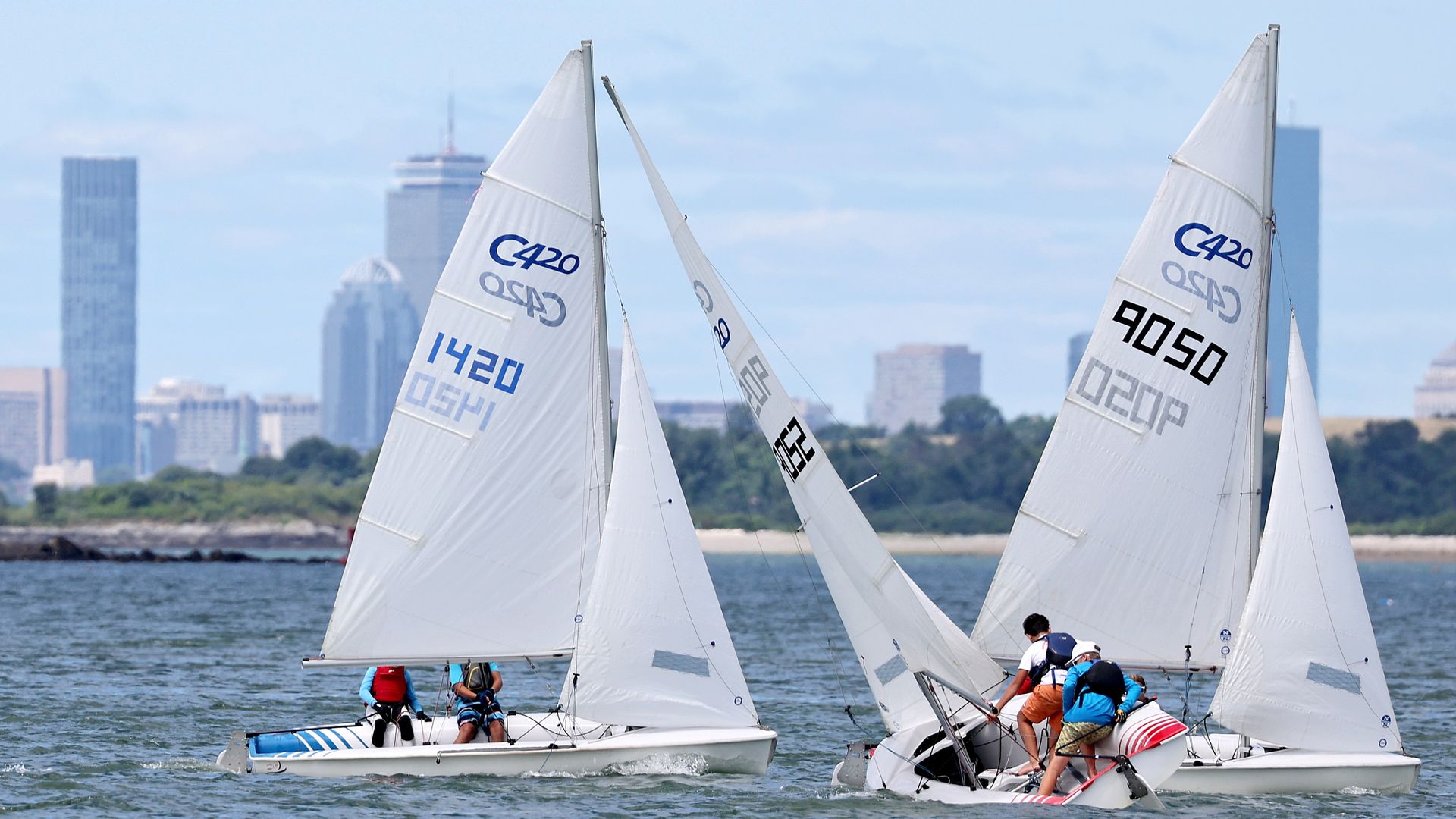 Two white sailboats with C420 markings and numbers 1420 and 9050 sailing on blue water near a shoreline with green trees and tall city buildings in the background under a partly cloudy sky.