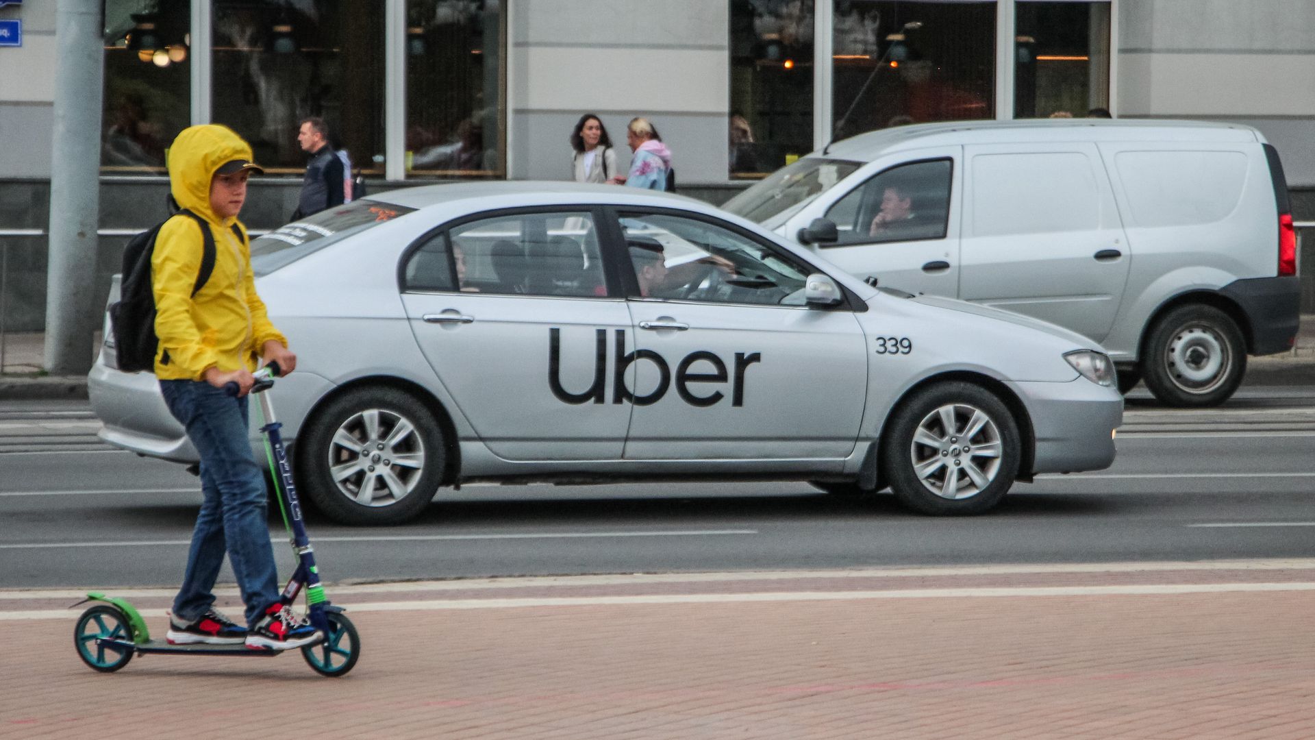 A child rides his scooter in the foreground as an uber car drives in the back.