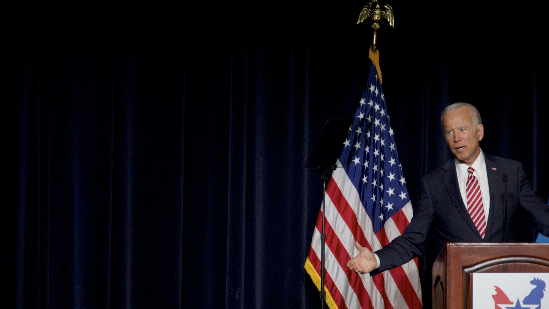 In this image, Biden gestures his arms outwards from behind a podium on stage. The American flag is behind him.