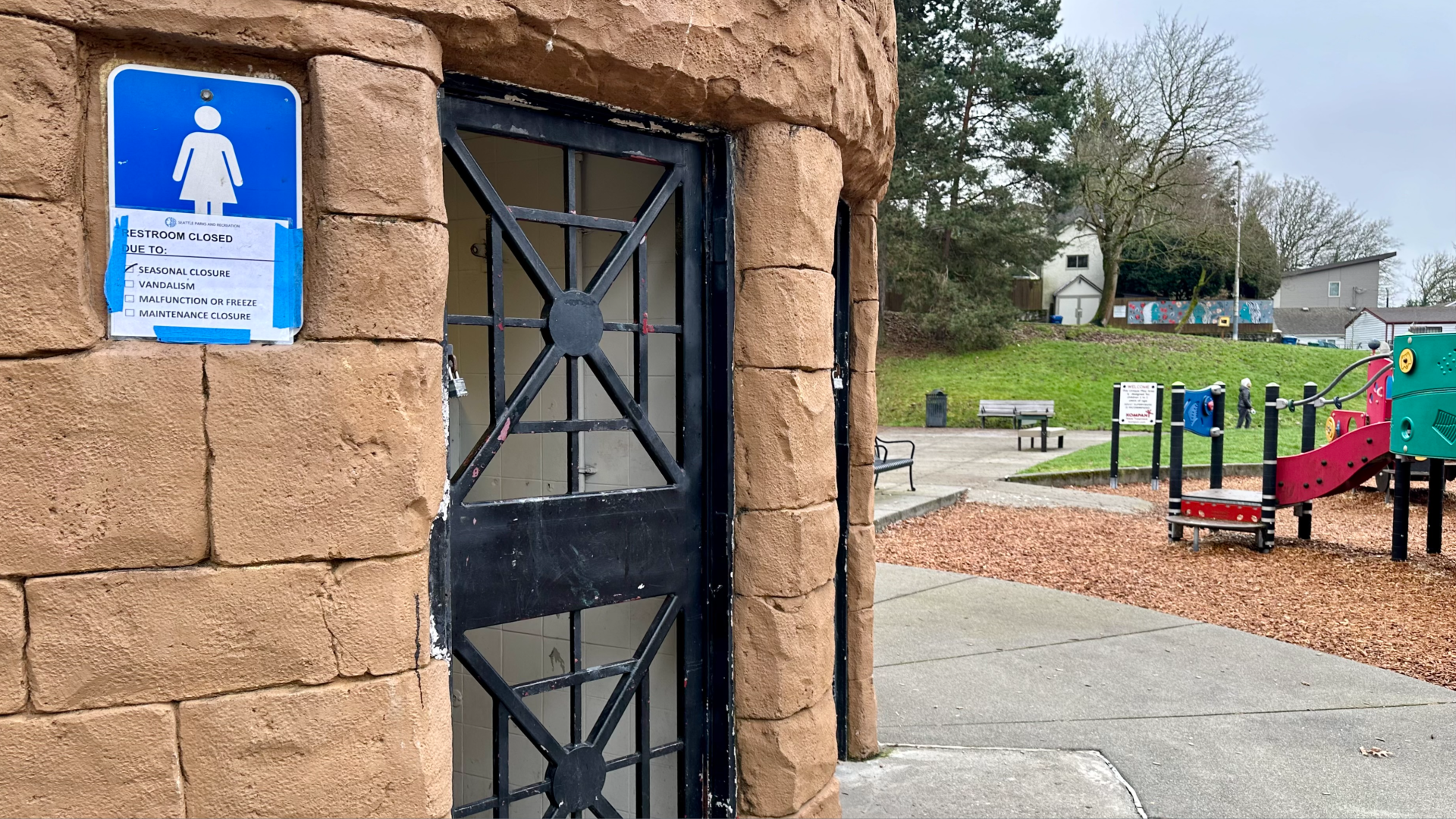 A park bathroom with a closure sign over the restroom sign, with a playground shown to the side.