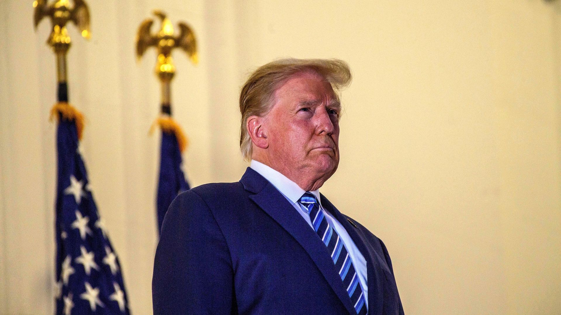 President Trump stands in front of two American flags while wearing a suit and blue striped tie