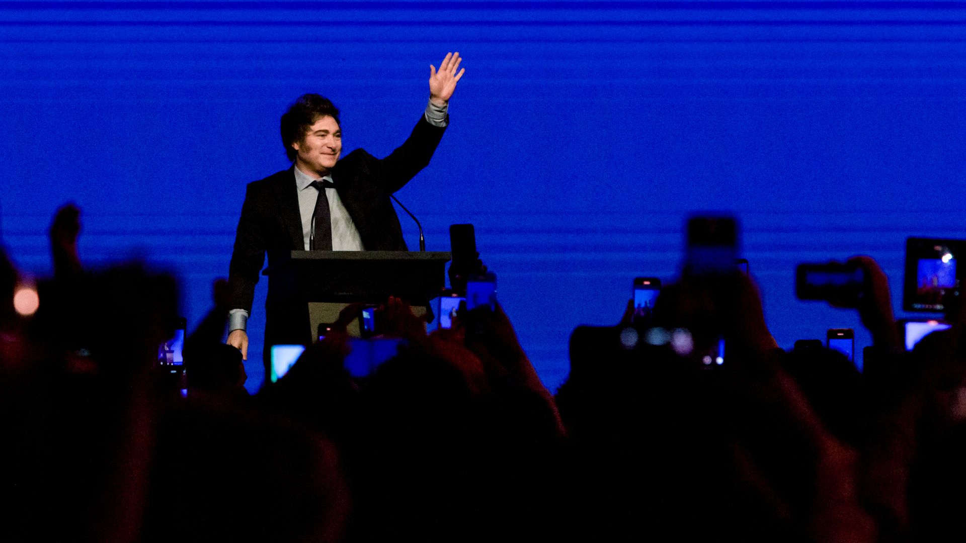 Argentina President Javier Milei waves to a crowd during a conference. There is a dark blue background behind him. 