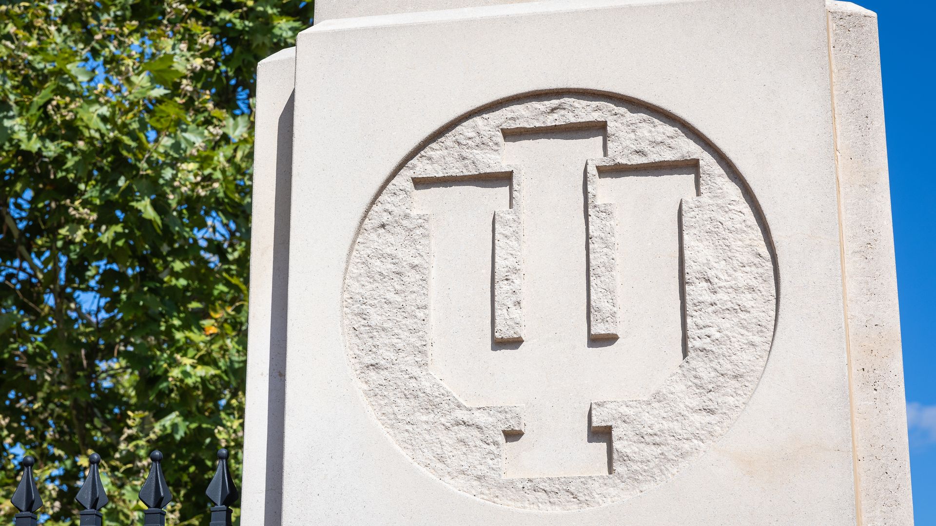 Stone monument with a carved circular emblem featuring an interlocking IU logo, set against a backdrop of green trees and blue sky.