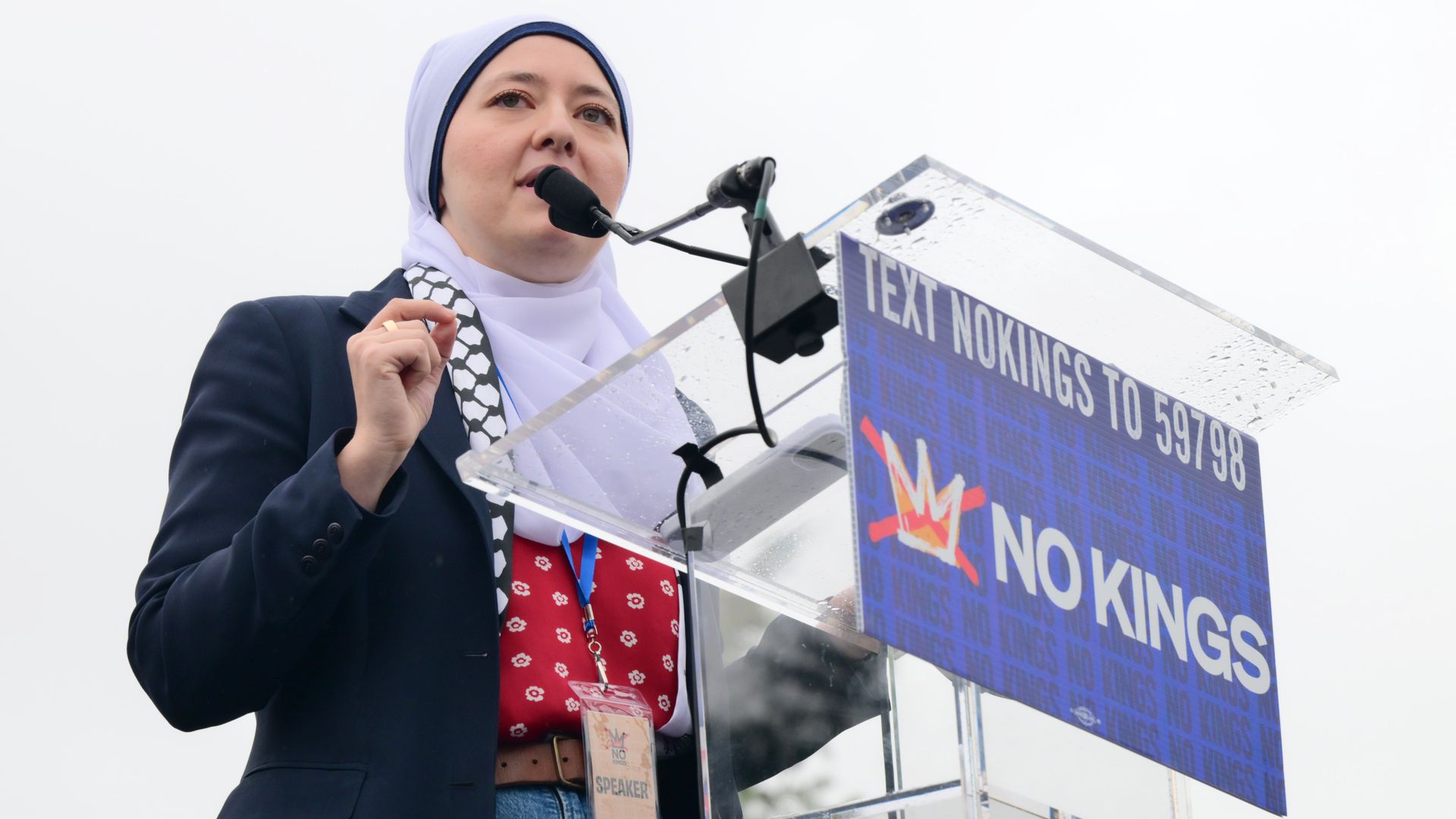 A person wearing a white headscarf and blue sports jacket speaks at a glass podium with a "No Kings" poster on its front.