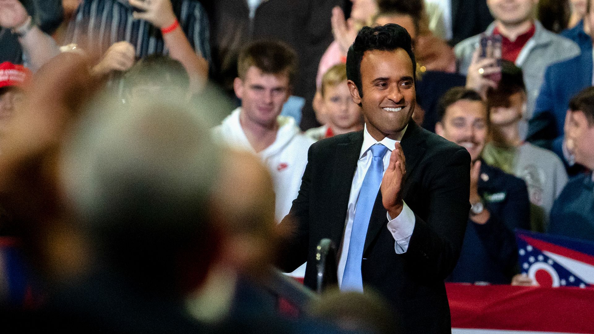 Indian man in black suit and blue tie claps at campaign event during speech he is presumably giving.