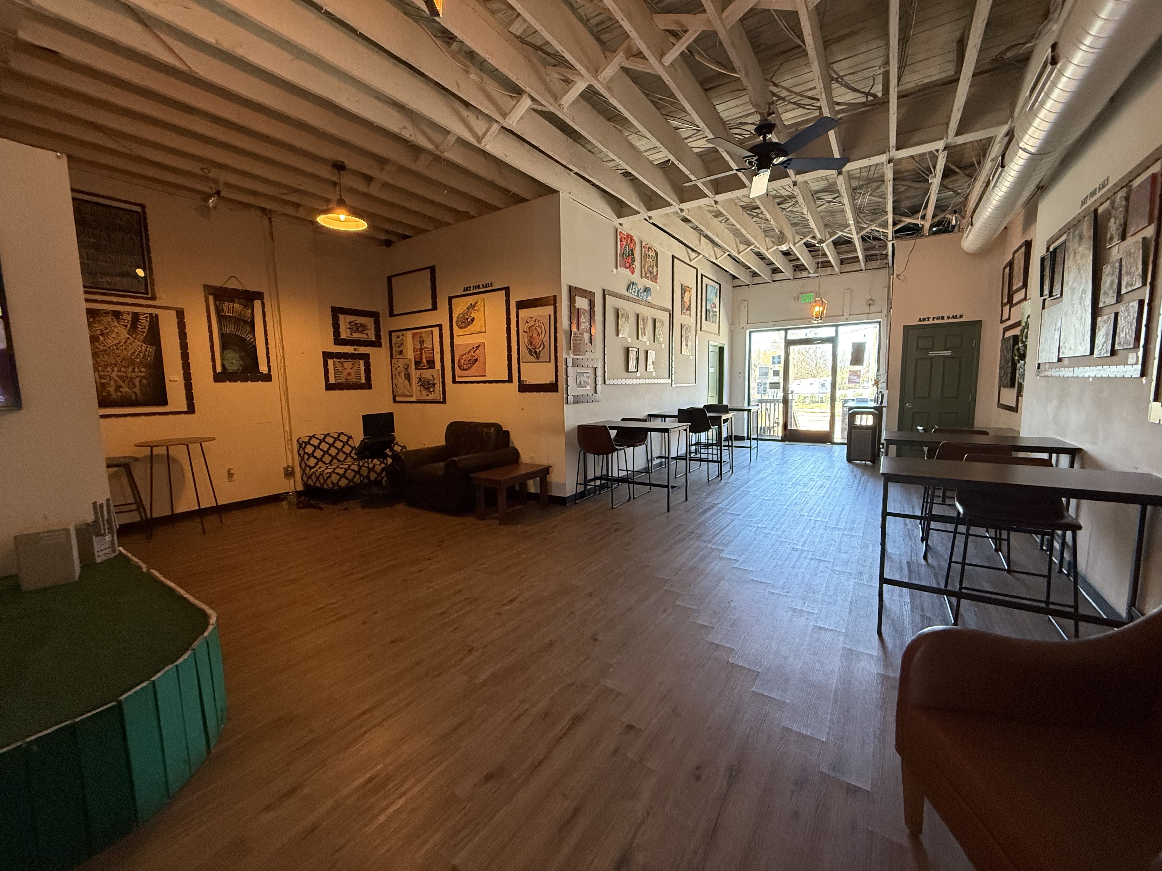 Wide view of an art space with exposed ceiling beams and a wooden floor. Left: seating area and small stage; right: tall tables with stools beside framed artworks. Door at far end lets in daylight.