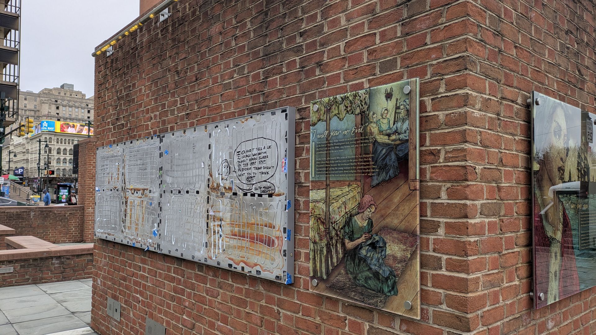 Red brick wall on a city street corner displaying historical and modern art plaques, including a comic-style speech bubble and an illustration of people inside a room.