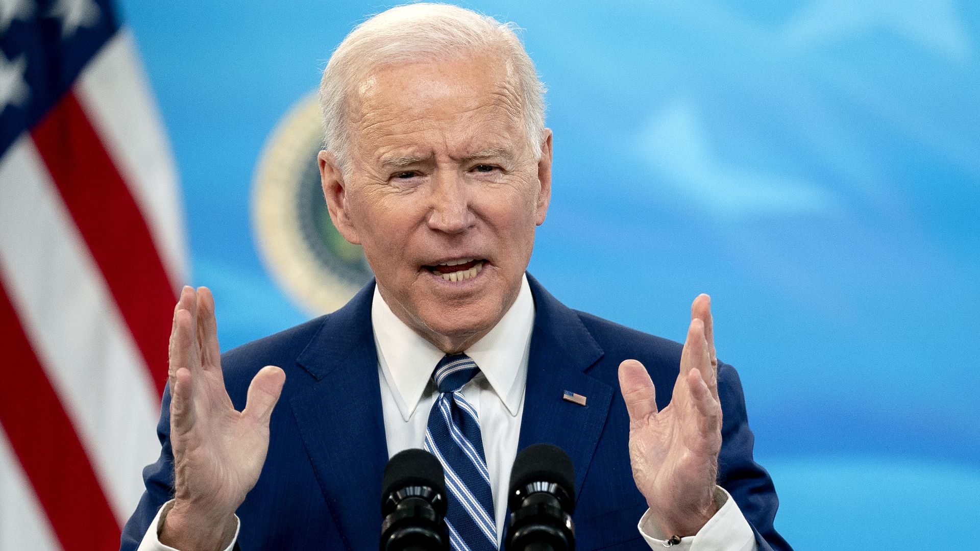 President Biden gestures with his hands while giving a speech with a flag and official seal in the background.