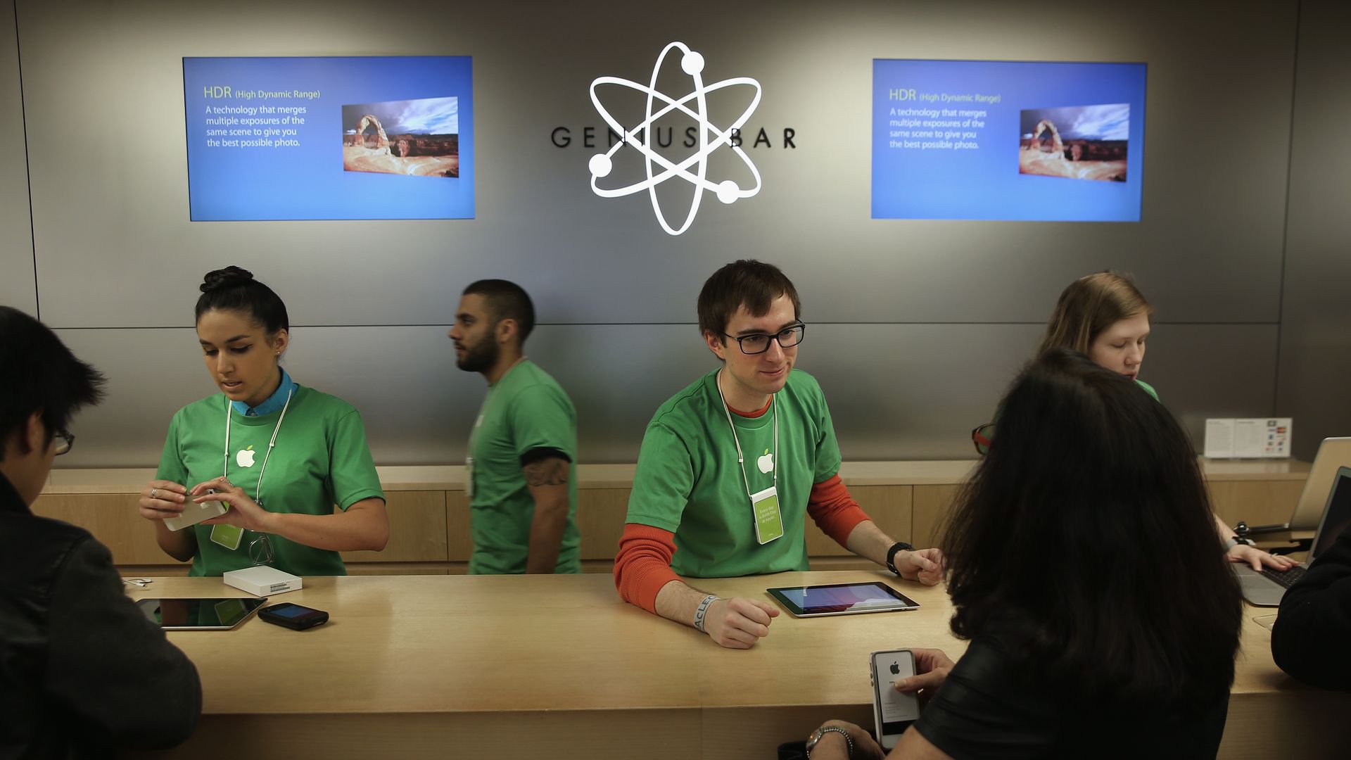 A man in a green shirt, layered over a red long-sleeved shirt, leans over a counter in front of a sign that says "Genius Bar"