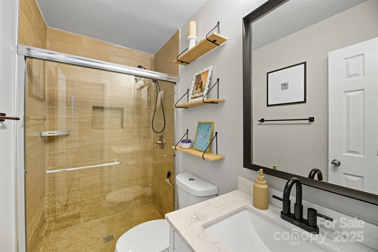 Modern bathroom with beige tiled walk-in shower with glass sliding door, white toilet, marble countertop, black faucet, beige soap dispenser, wooden wall shelves with decor, and large mirror.