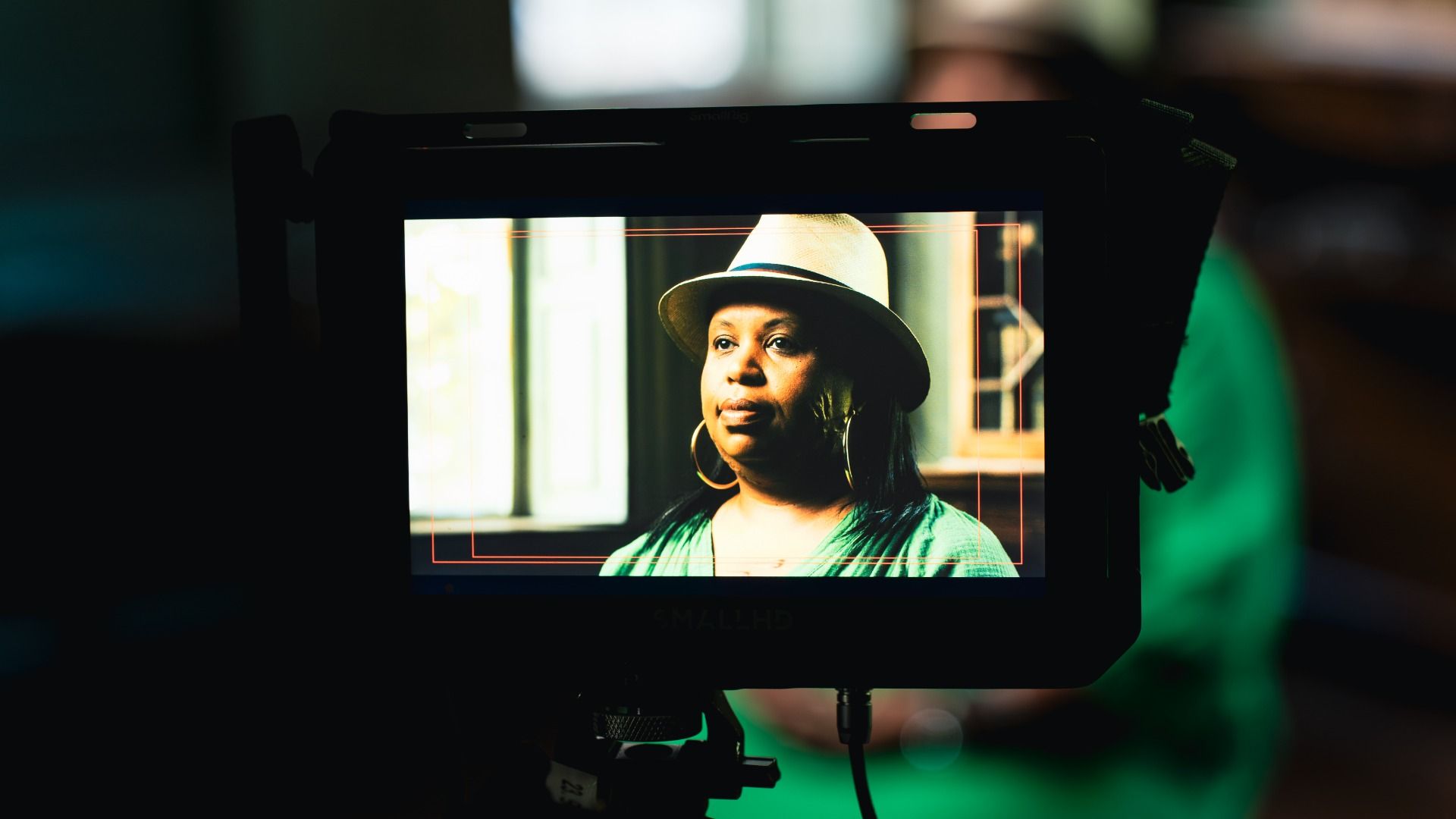 On a camera monitor, a woman wearing a light hat and large hoop earrings gazes off-camera. Warm light, red framing lines outline her portrait against a dark studio backdrop.