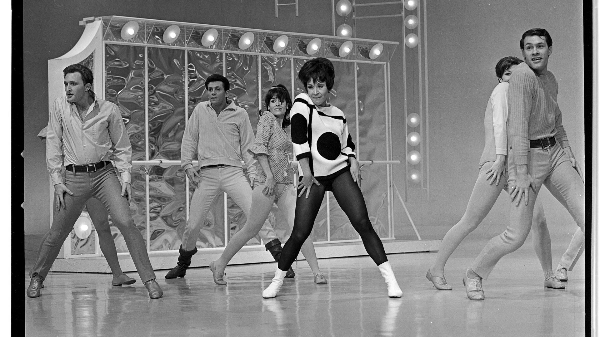 Several men dancers surround Chita Rivera as she dances on a stage with lights behind her