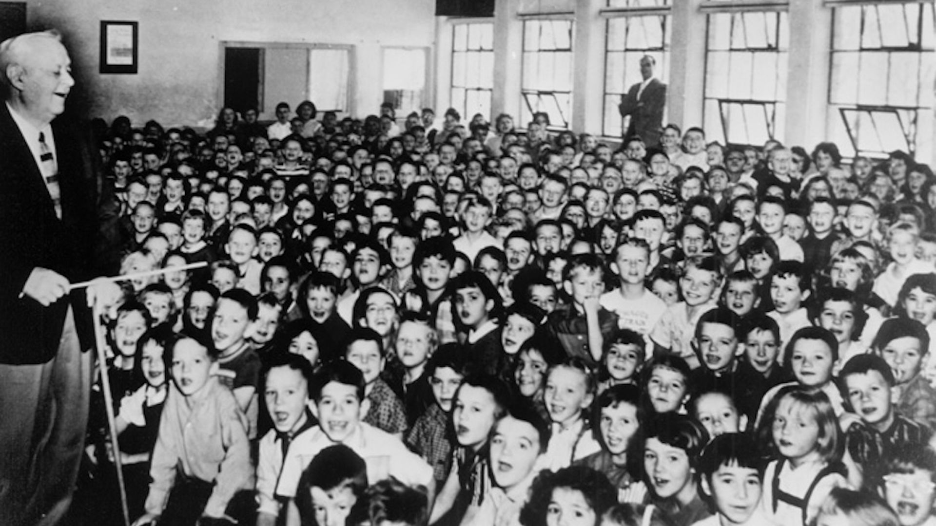 A black and white photo of a man conducting  a gymnasium full of children singing