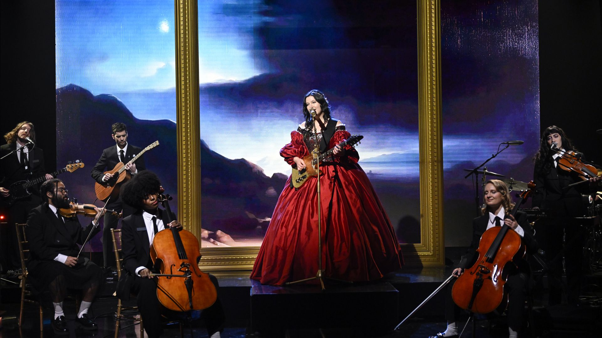 An artist in a big red dress in front of a giant empty portrait on stage while surrounded by an orchestra