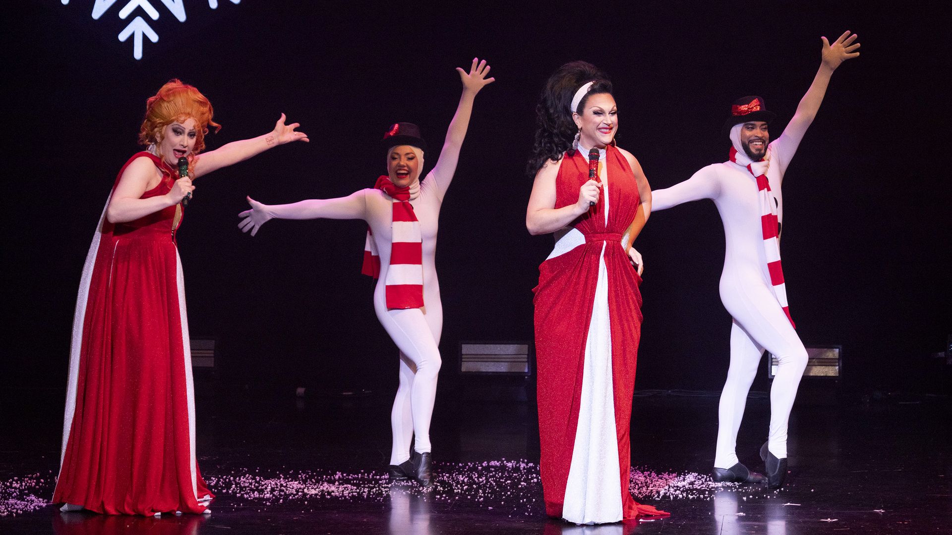 Four performers on stage in festive holiday costumes: two in red and white dresses with microphones, and two in white bodysuits with red scarves and hats, under a large white snowflake.