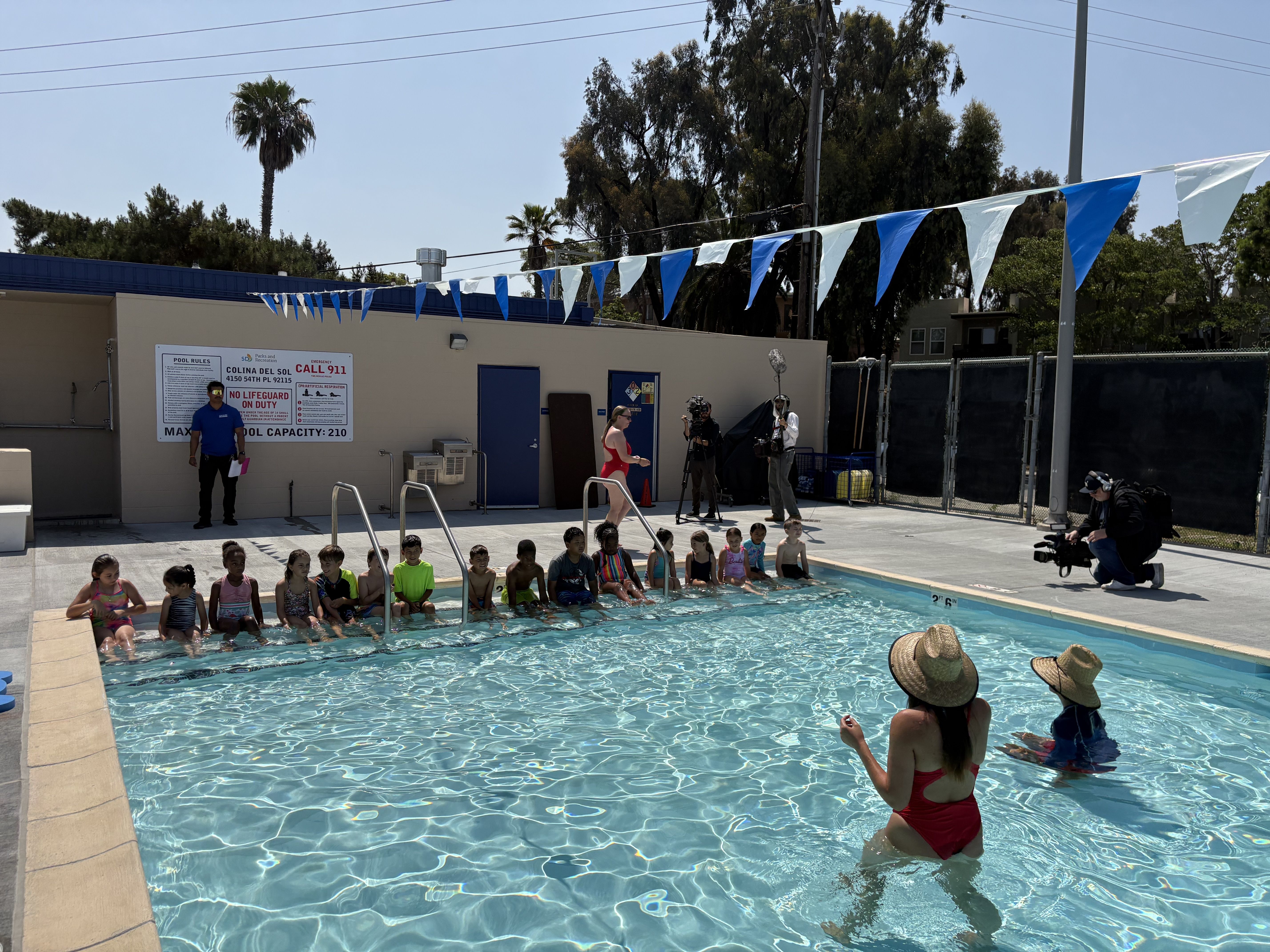 Children sit along the edge of a sunny outdoor pool, with a few adults and photographers nearby. Blue and white pennants hang above, palm trees and a beige building in the background.
