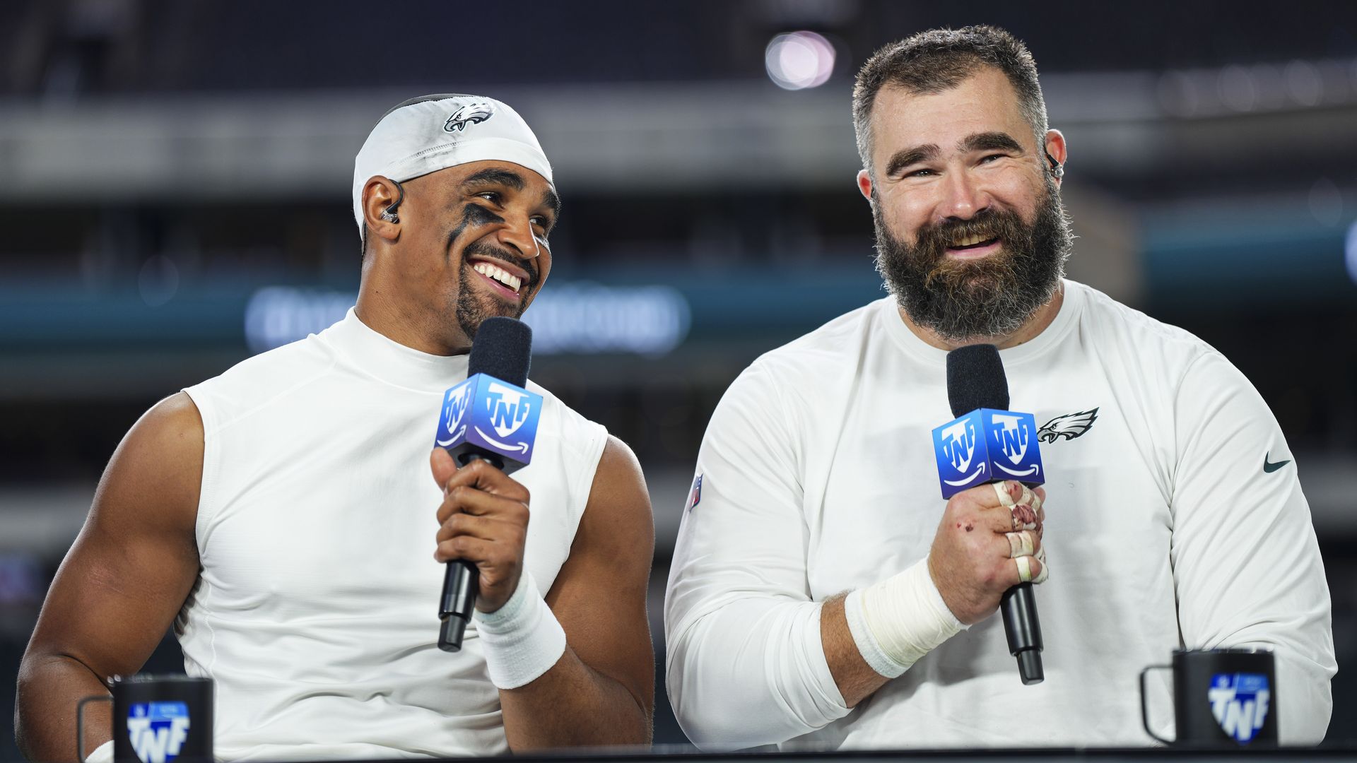 Jalen Hurts smiles at Jason Kelce while they both hold up microphones during a postgame broadcast.