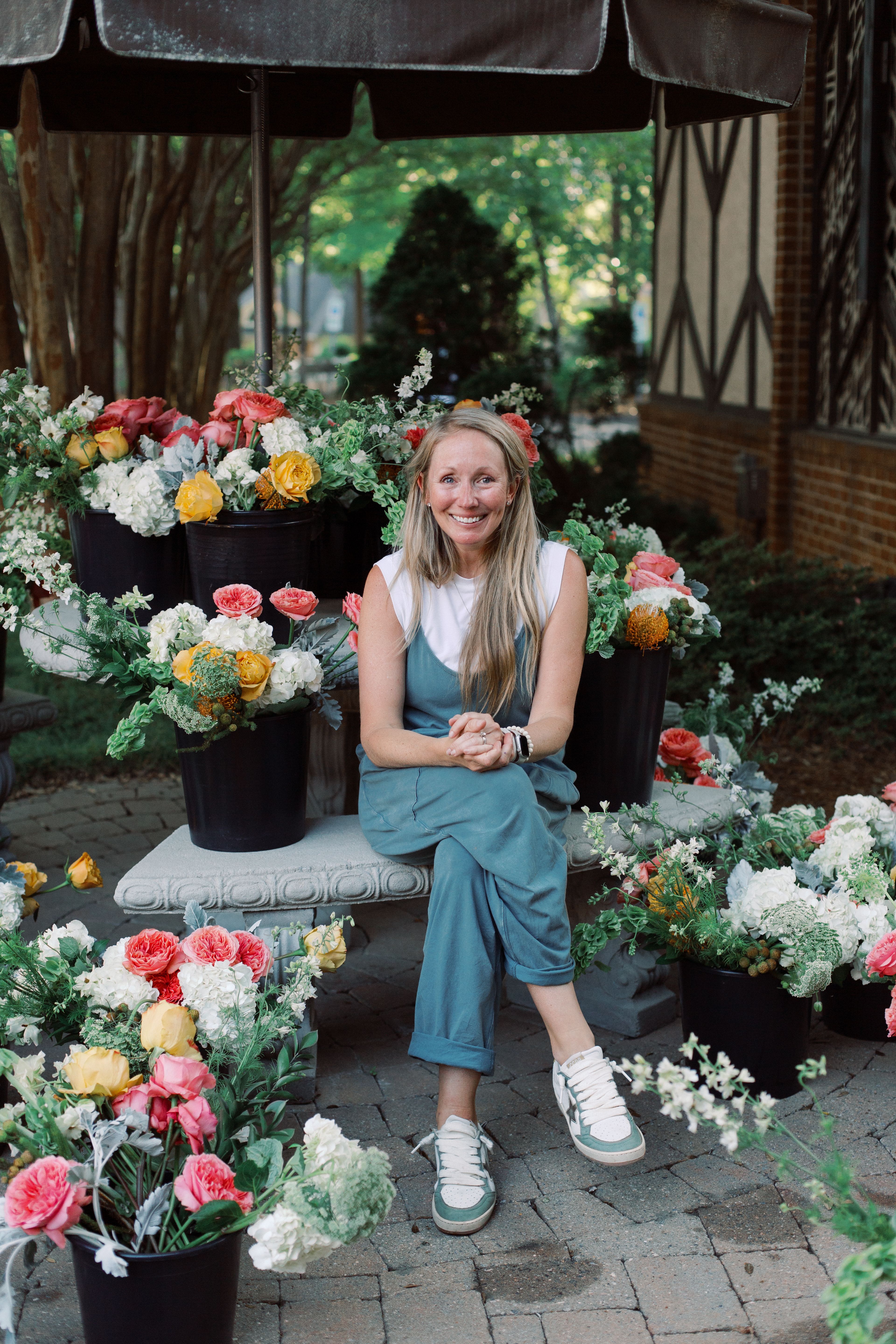 A smiling woman sits on a stone bench among vibrant pink, white, and yellow flowers in black buckets, under a large umbrella, outdoors with trees and a brick building nearby.