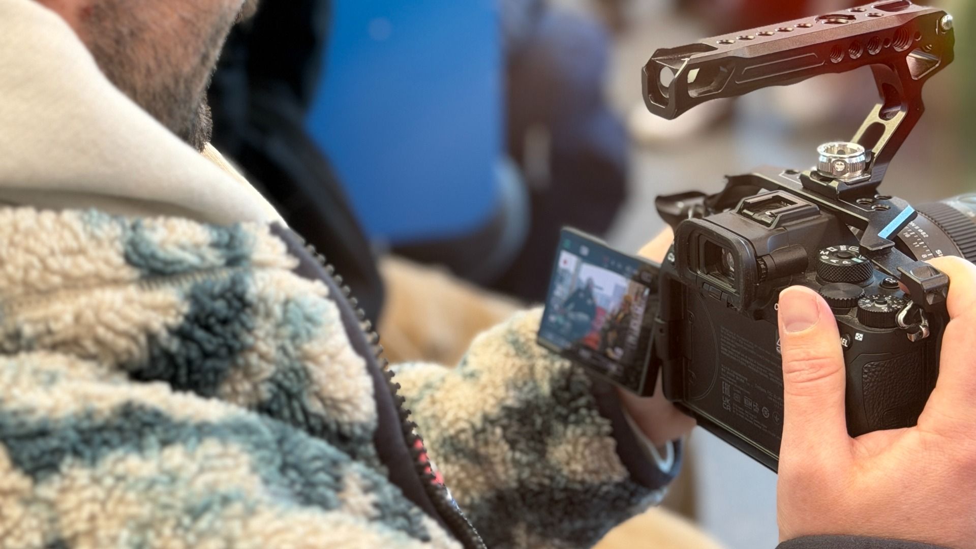 Close-up of a person wearing a fuzzy blue and white patterned jacket holding a black camera with a flip-out screen showing two people in the frame.