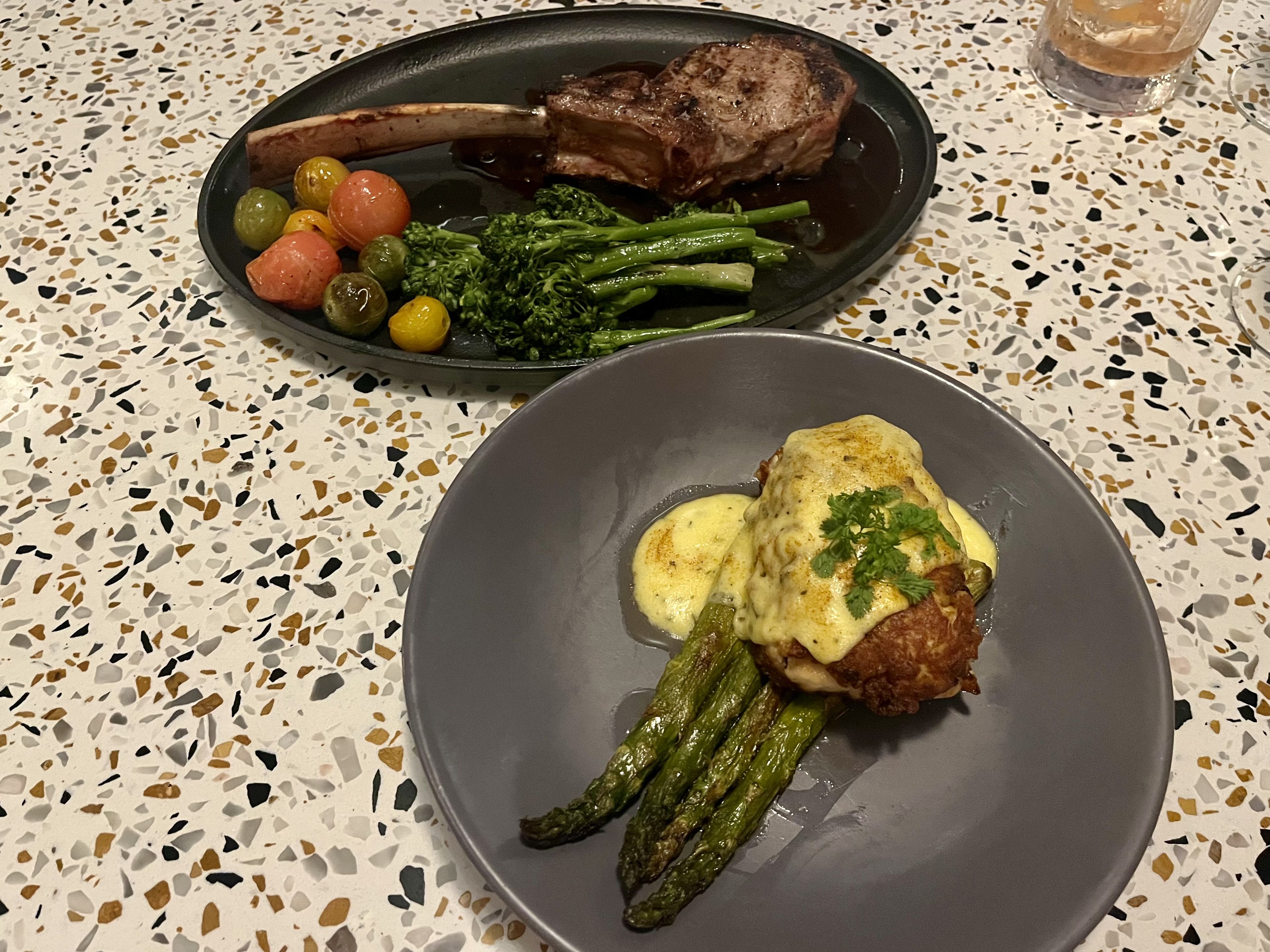 Two dark gray plates of food, one with a fried hunk of meat topped with yellow sauce on top of a bed of asparagus, another with a large steak with the bone still in next to a bunch of small red and green tomatoes and a bunch of broccolini. 