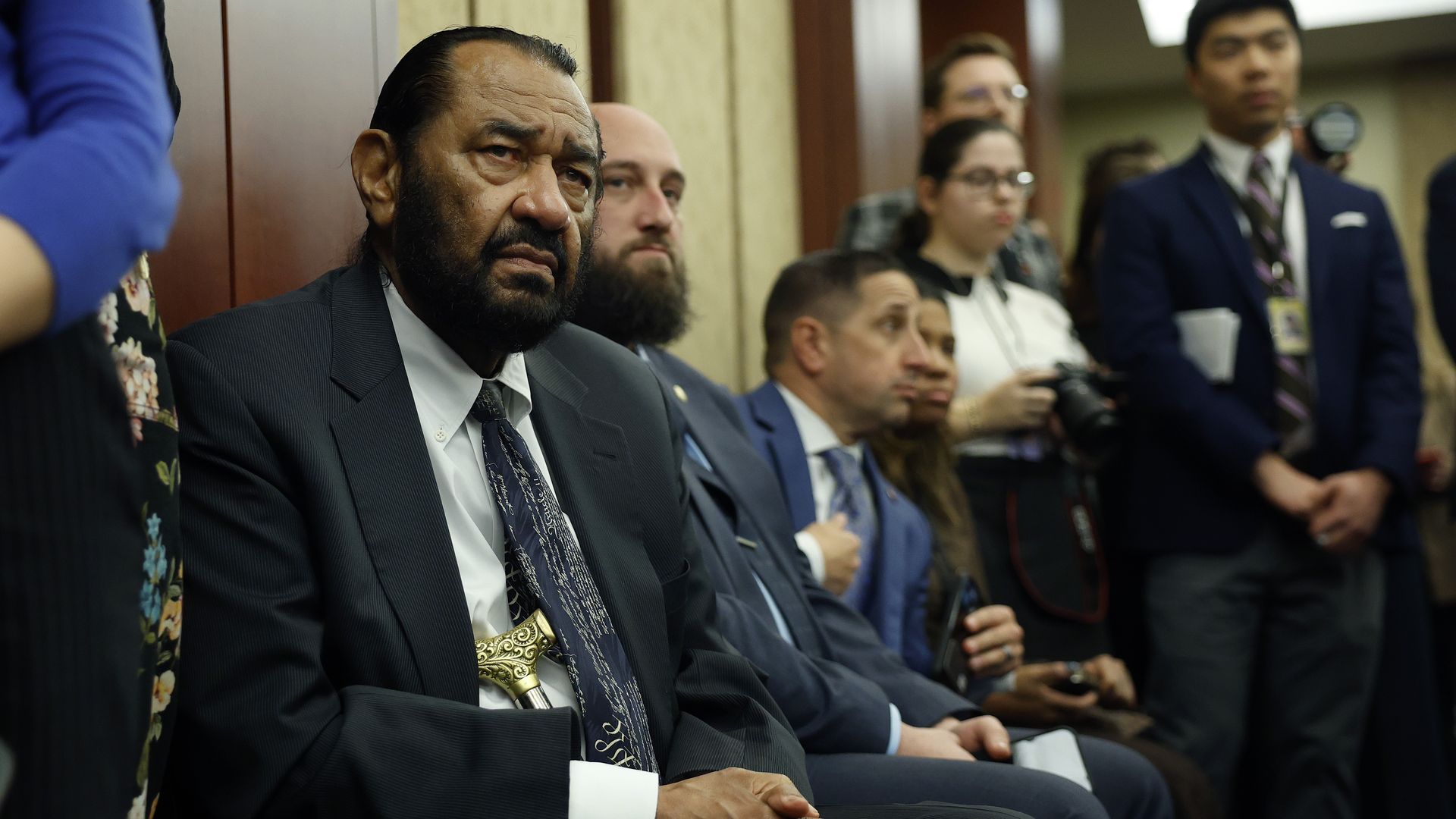 Rep. Al Green sits in a wood-paneled room while flanked by reporters, staff and security.