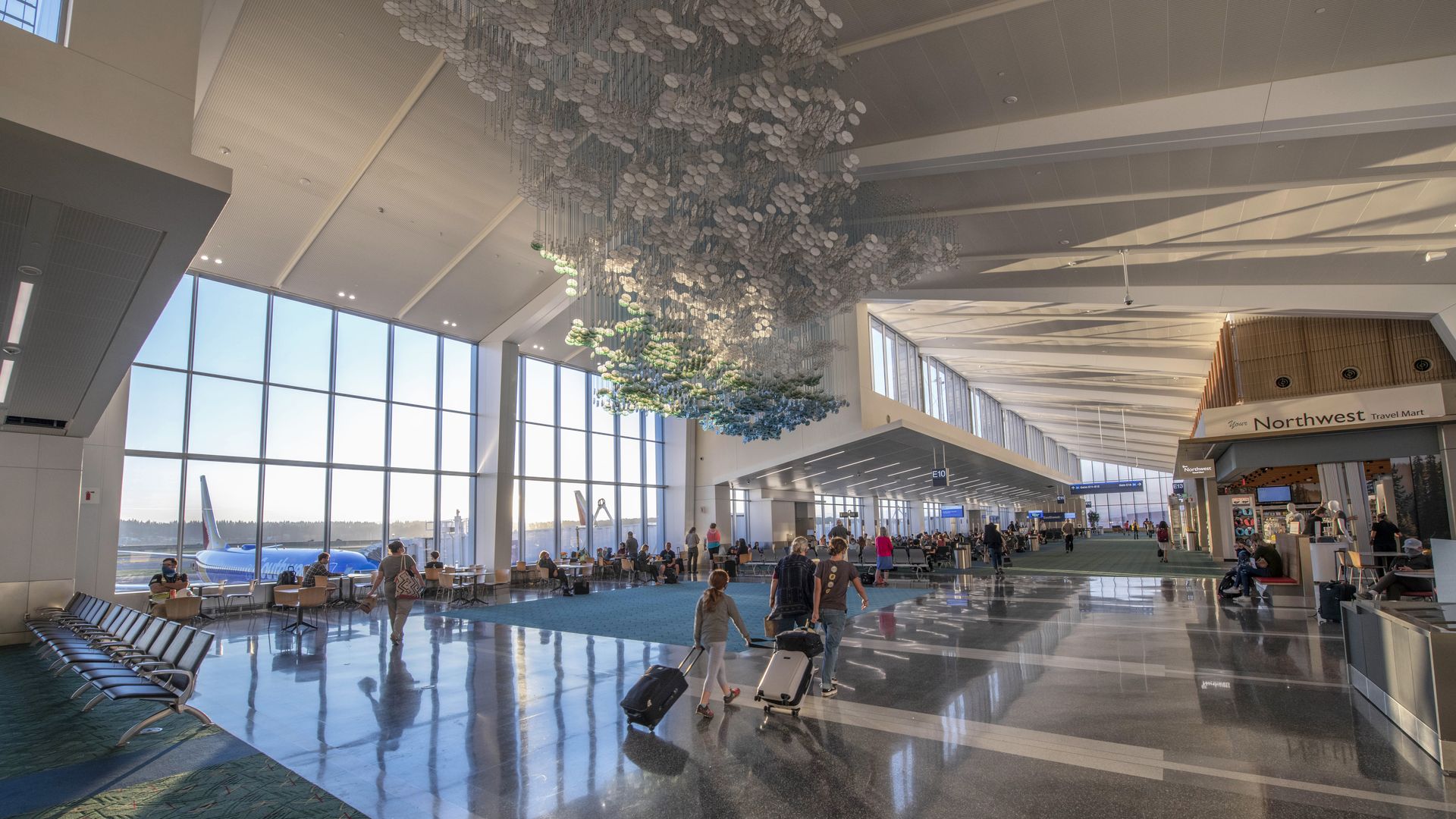 Busy airport terminal with large windows showing a blue airplane outside, passengers walking with suitcases, a hanging art installation, and a travel mart kiosk on the right.