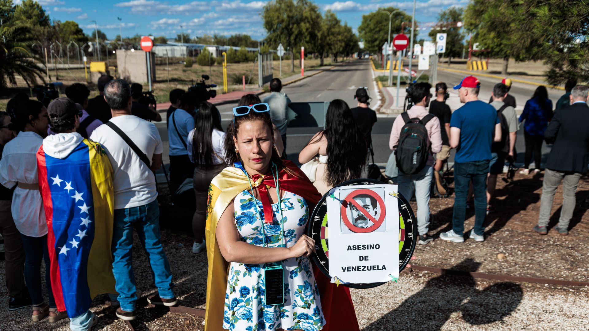 A group of people waiting for Venezuelan exile Edmundo Gonzalez are shown from behind, except for a woman facing the camera who is wearing a Venezuela flag like a cape and holding a white paper sign that has a crossed out picture of Nicolas Maduro and says "murderer of Venezuela" in Spanish
