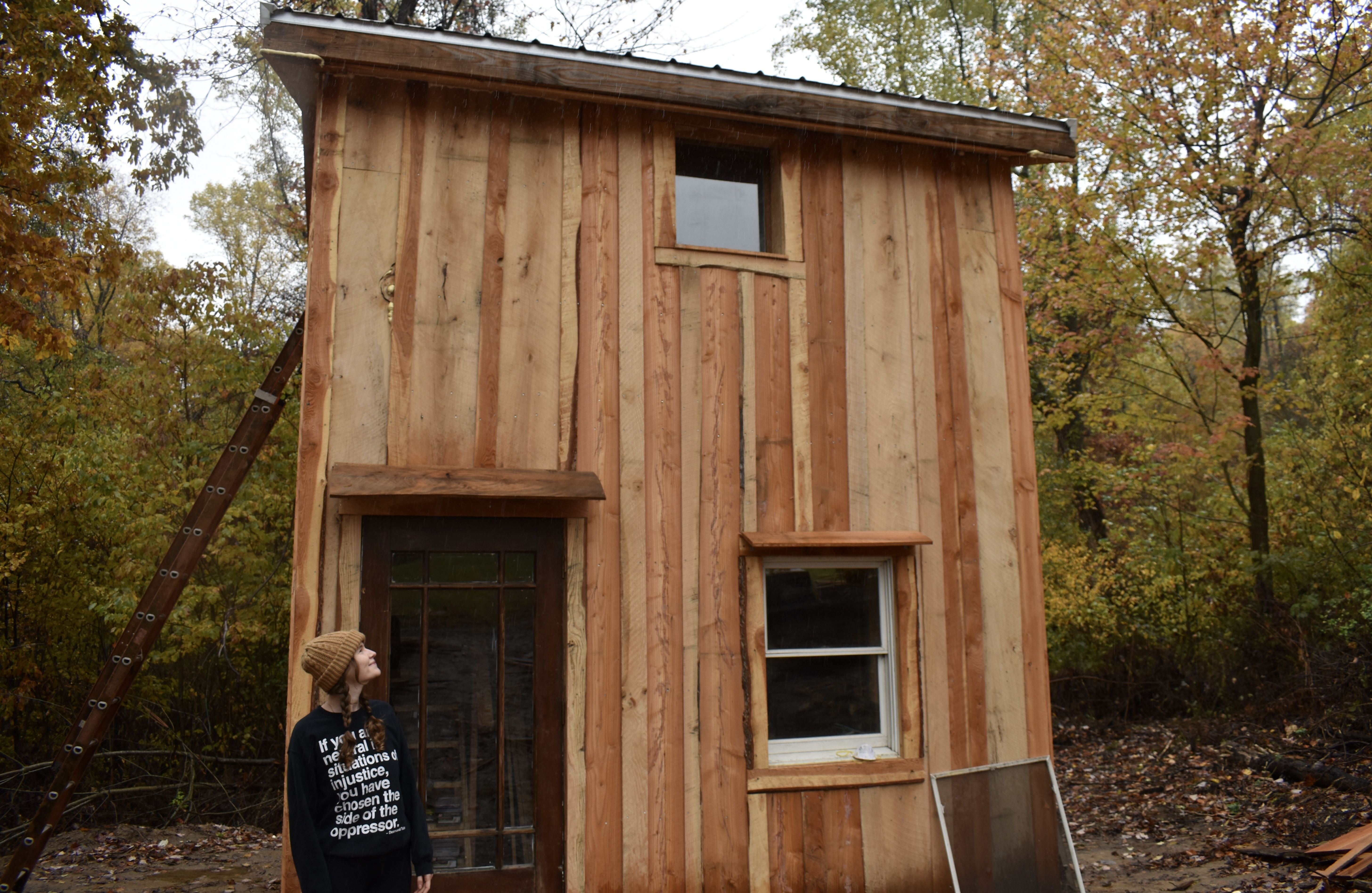 Person in a beanie and black sweater stands beside a small wooden cabin with two windows and a door, surrounded by autumn-colored trees in a forest setting.