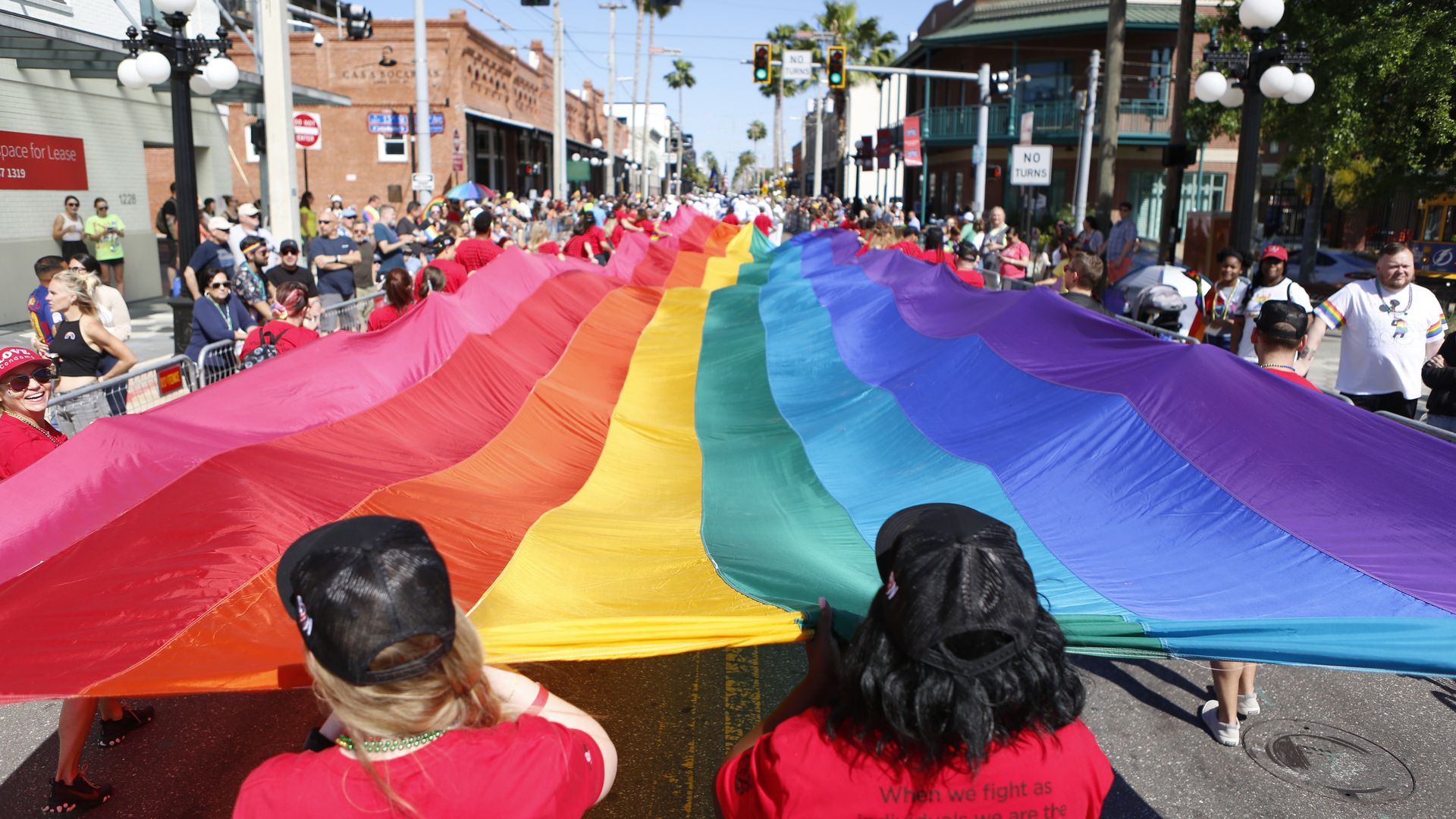 People hold a large rainbow pride flag stretched across a sunny city street during a vibrant Pride parade with onlookers and historic buildings.